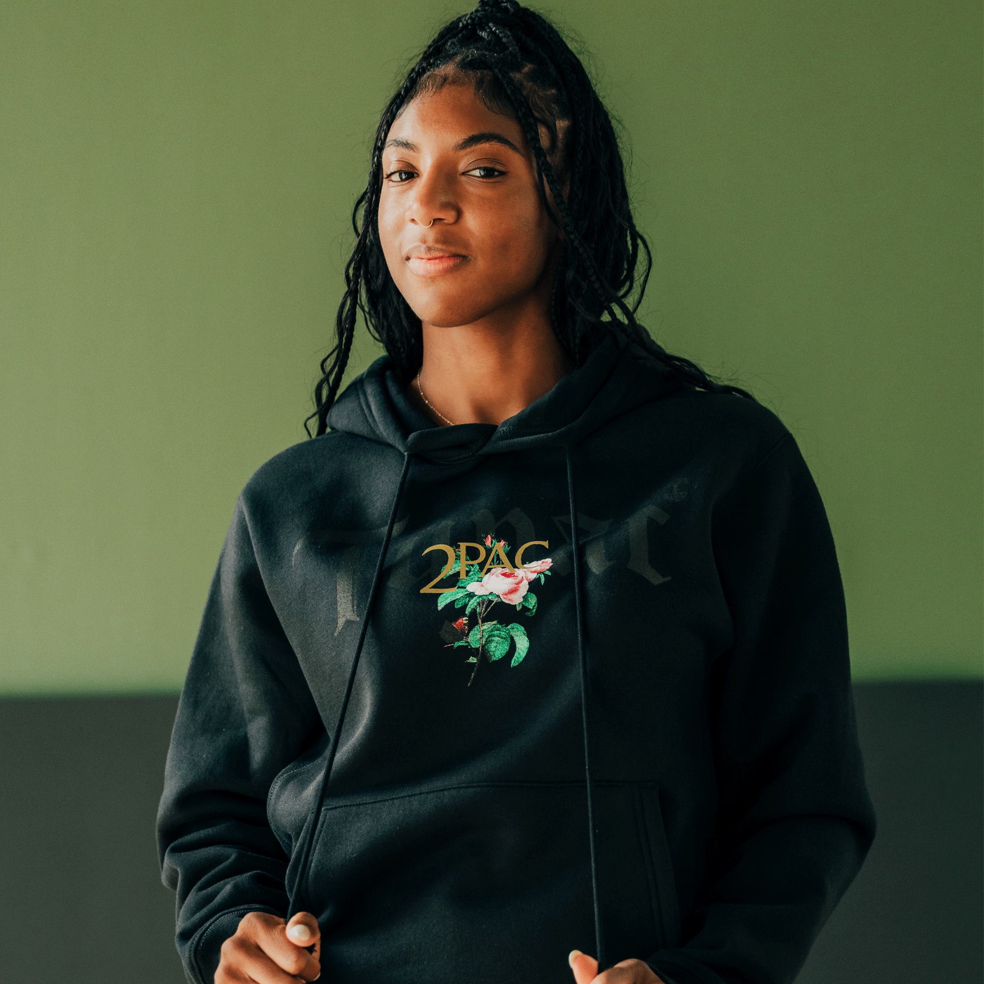 A person wearing the Roses Hoodie from Oaklandish stands confidently against a two-tone background. This piece, part of the exclusive Oaklandish x Tupac hip-hop collection, features a floral design and "2PAC" on the front. They have long braided hair and hold the hoodie strings, while soft lighting highlights their relaxed expression.