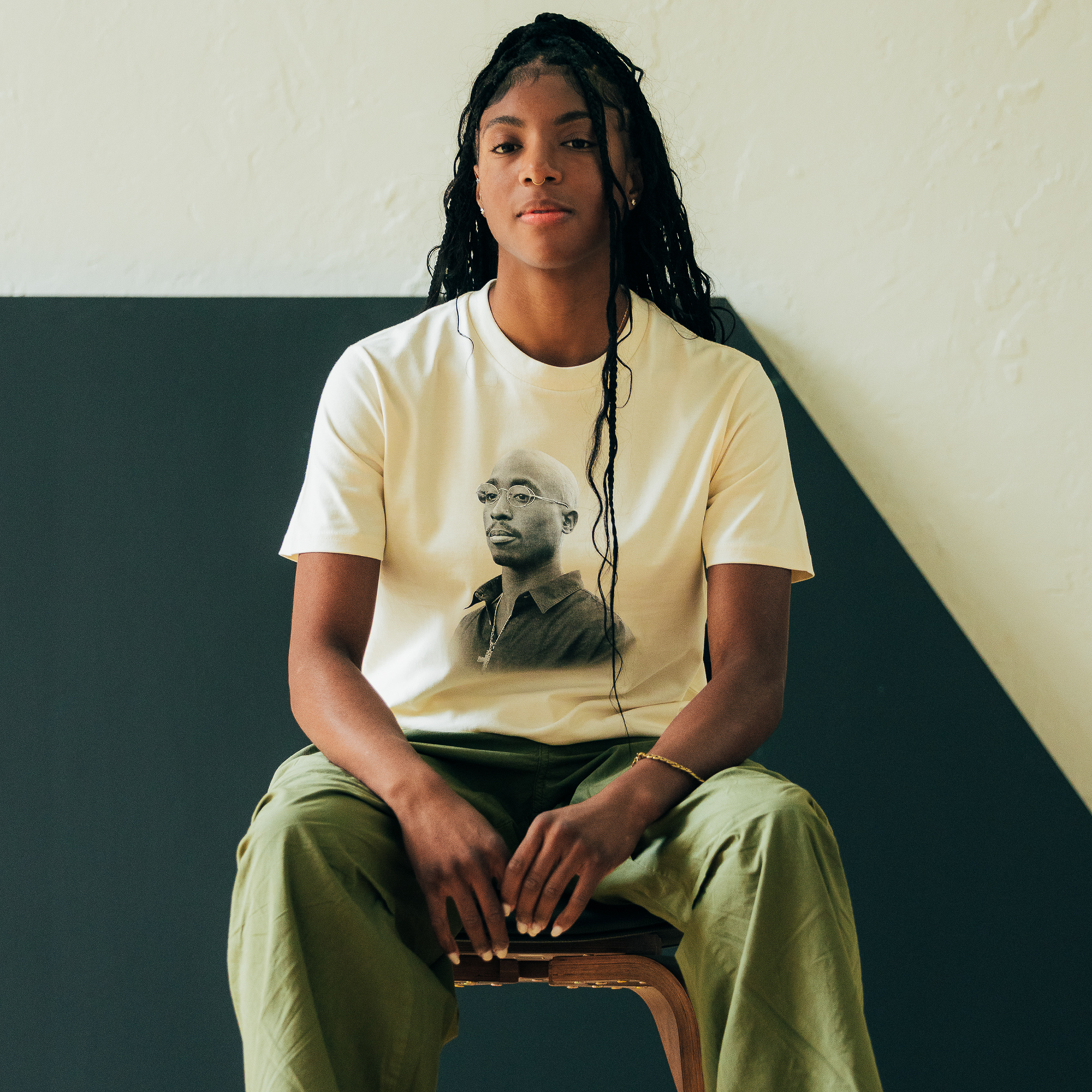 A person with long braided hair sits on a stool, wearing the Sometimes Eye Cry Tee by Oaklandish—a white T-shirt showcasing a grayscale portrait of Tupac Shakur paired with green pants. The background, inspired by Oaklandish style, is divided diagonally into light and dark sections, creating a minimalist contrast. They gaze directly at the camera with a calm demeanor.