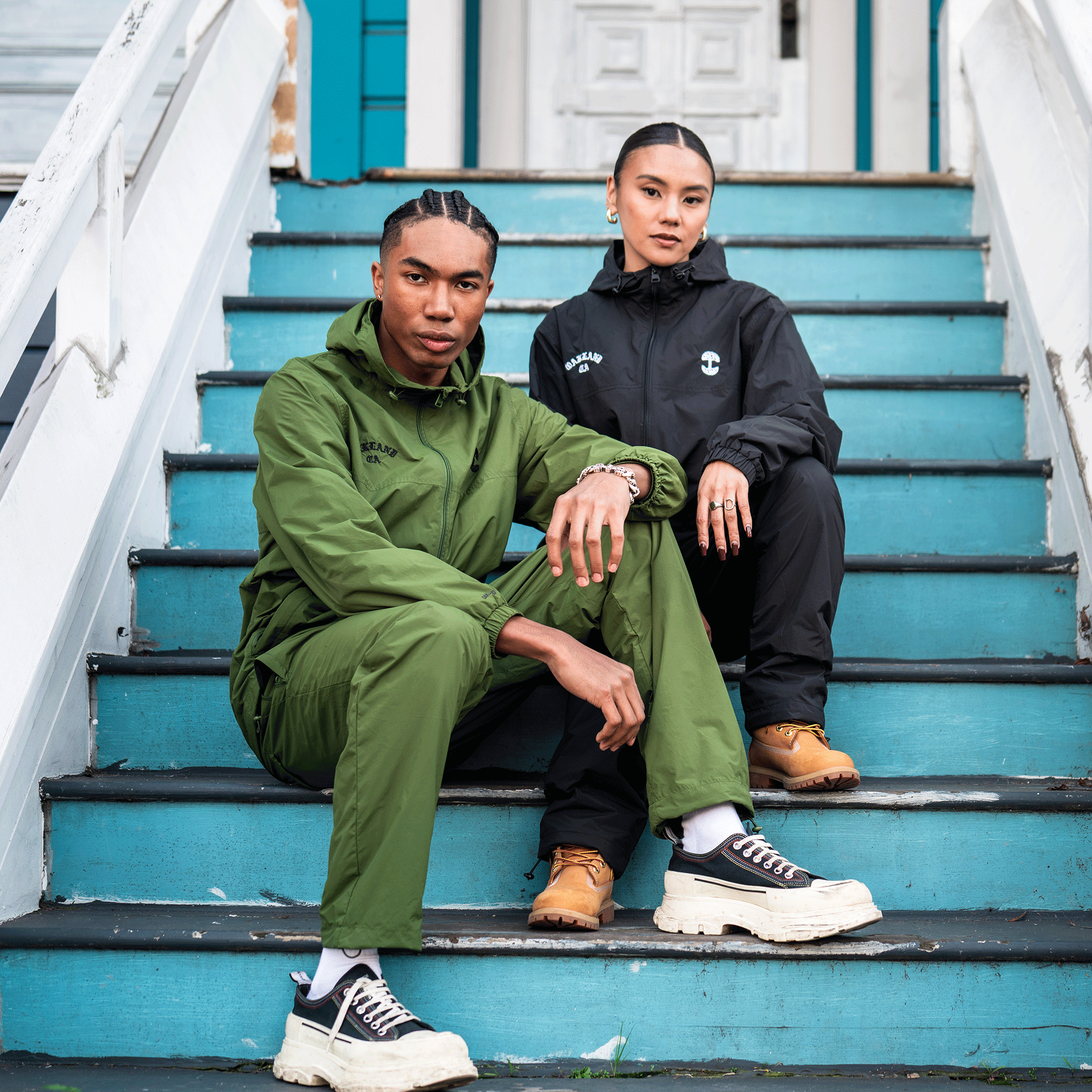 Two people sit side by side on turquoise steps. The person on the left wears a green tracksuit; the person on the right sports Oaklandish's Susurrus Track Top, black track bottoms, and tan boots. Both pose confidently for the camera.