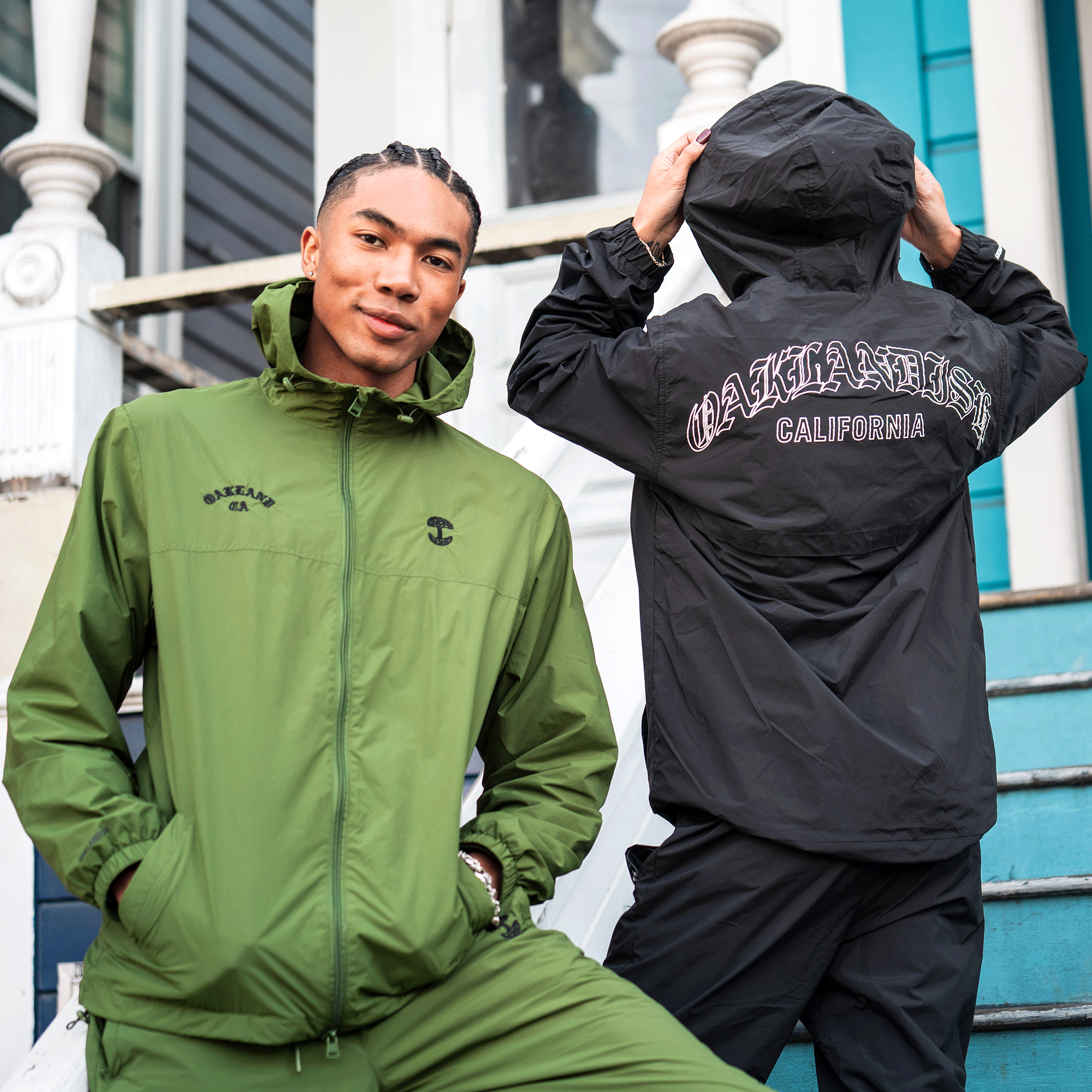 Two people pose outdoors. One faces forward in a green hooded nylon jacket and pants. The other wears the Oaklandish Susurrus Track Top in black with "OAKLANDISH CALIFORNIA" on the back. Blue and white houses complete the scene.