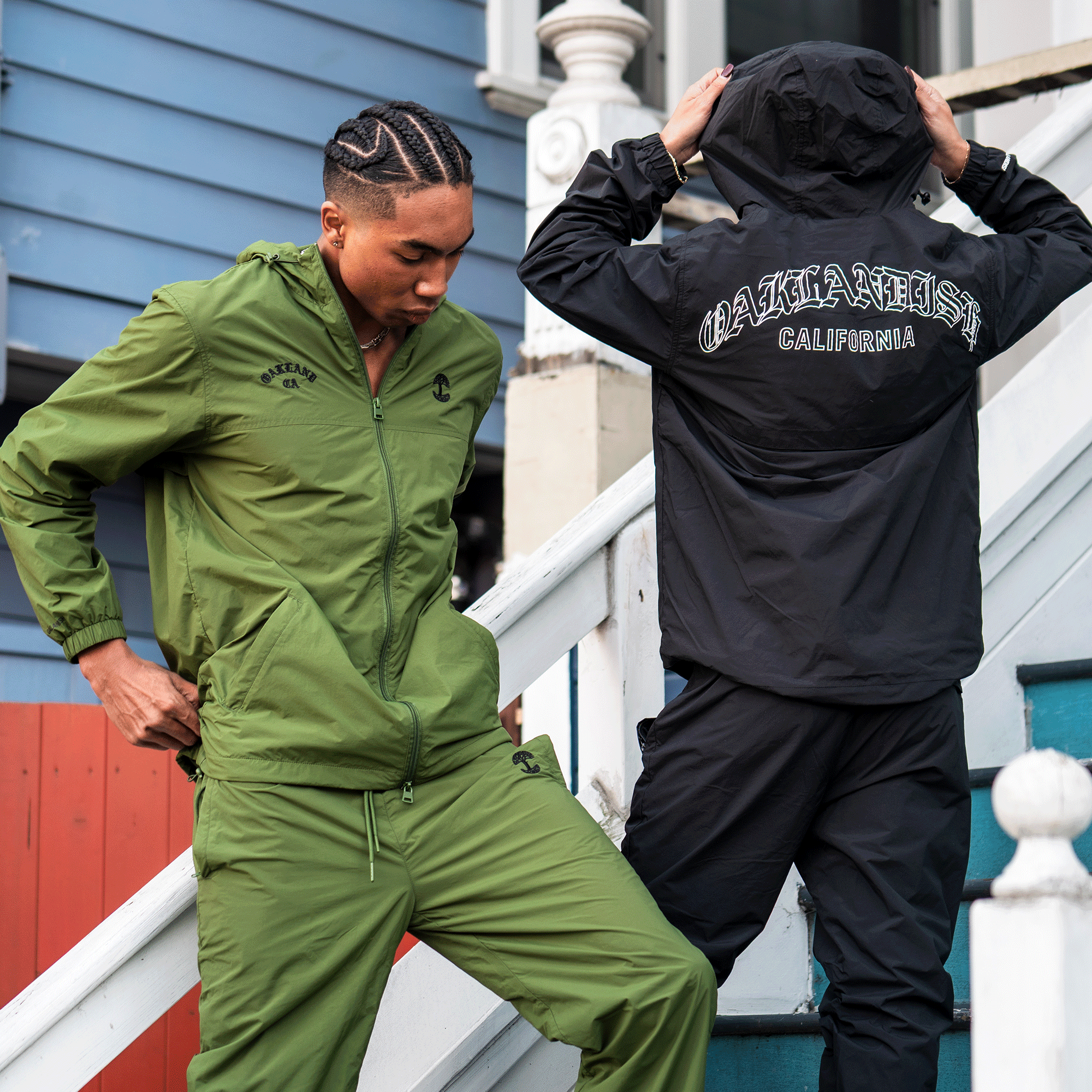 Two people in tracksuits stand on outdoor stairs. The left wears Oaklandish Susurrus Track Bottoms in olive green with an adjustable waistband. The right sports a black Oaklandish tracksuit with “OAKLANDISH CALIFORNIA” and a tree logo.