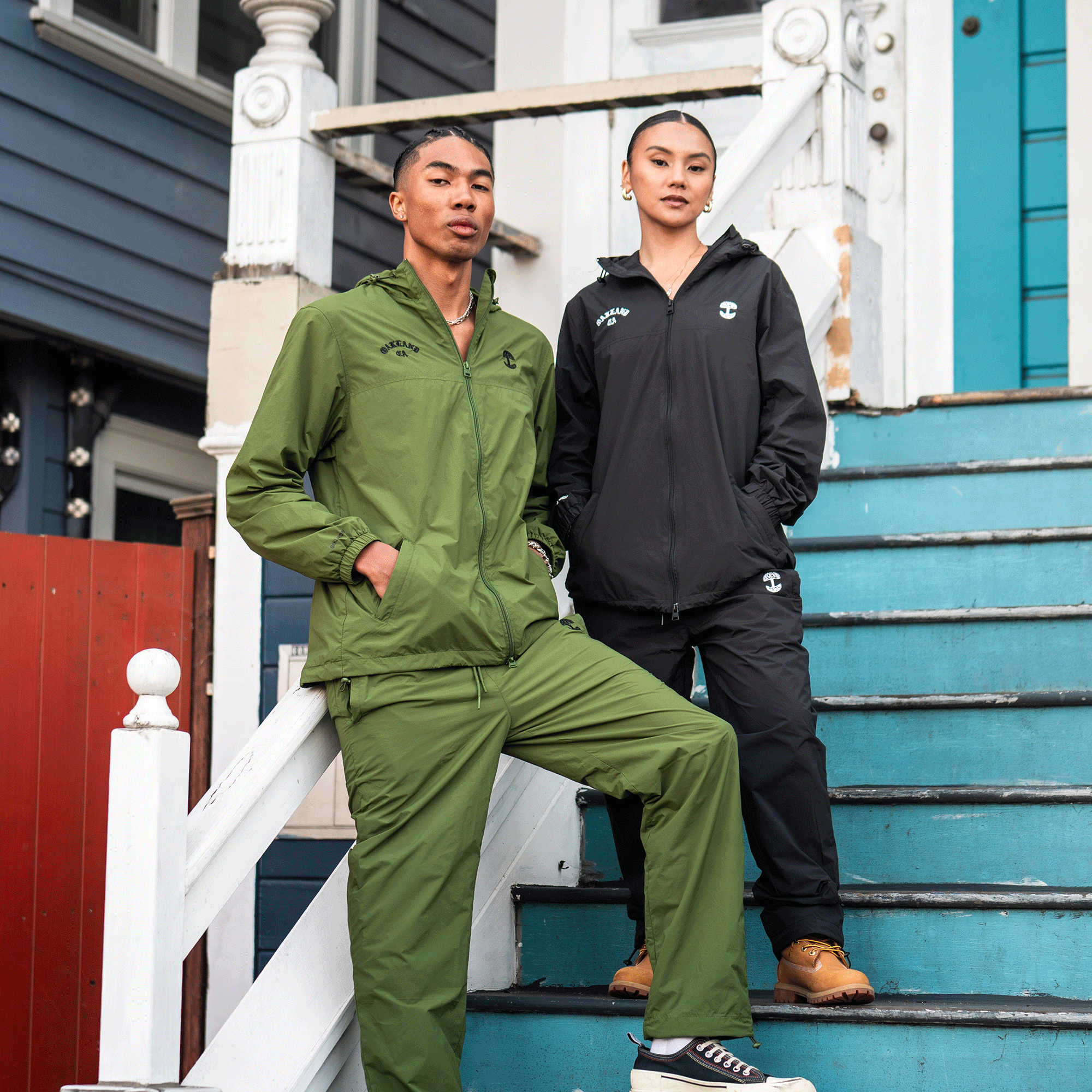 Two people pose on blue stairs outside a house. The person on the left wears a green Susurrus Track Bottom by Oaklandish with an adjustable waistband and white sneakers; the other, in black pants and tan boots, stands confidently one step down.