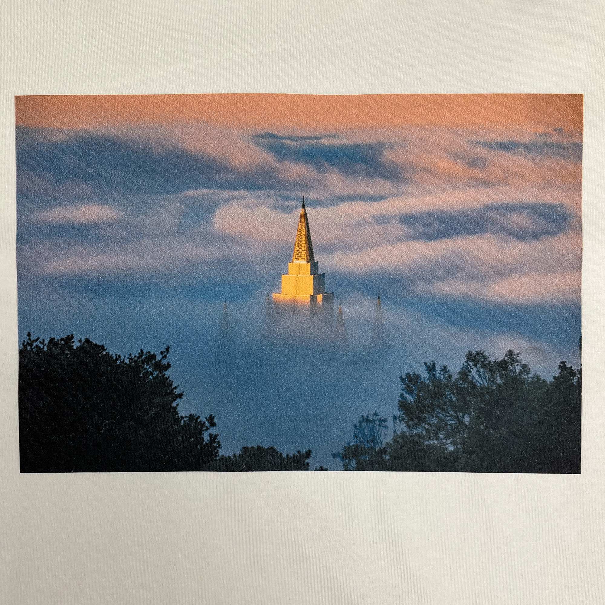 An image on the Temple Swirl Tee by Oaklandish shows an LDS temple with a pointed golden spire emerging above dense, rolling clouds at sunrise or sunset in the Oakland Hills. The sky is adorned with a gradient of pink and blue hues, while silhouettes of trees elegantly frame the bottom of the scene. The spire is beautifully illuminated in soft, warm light.