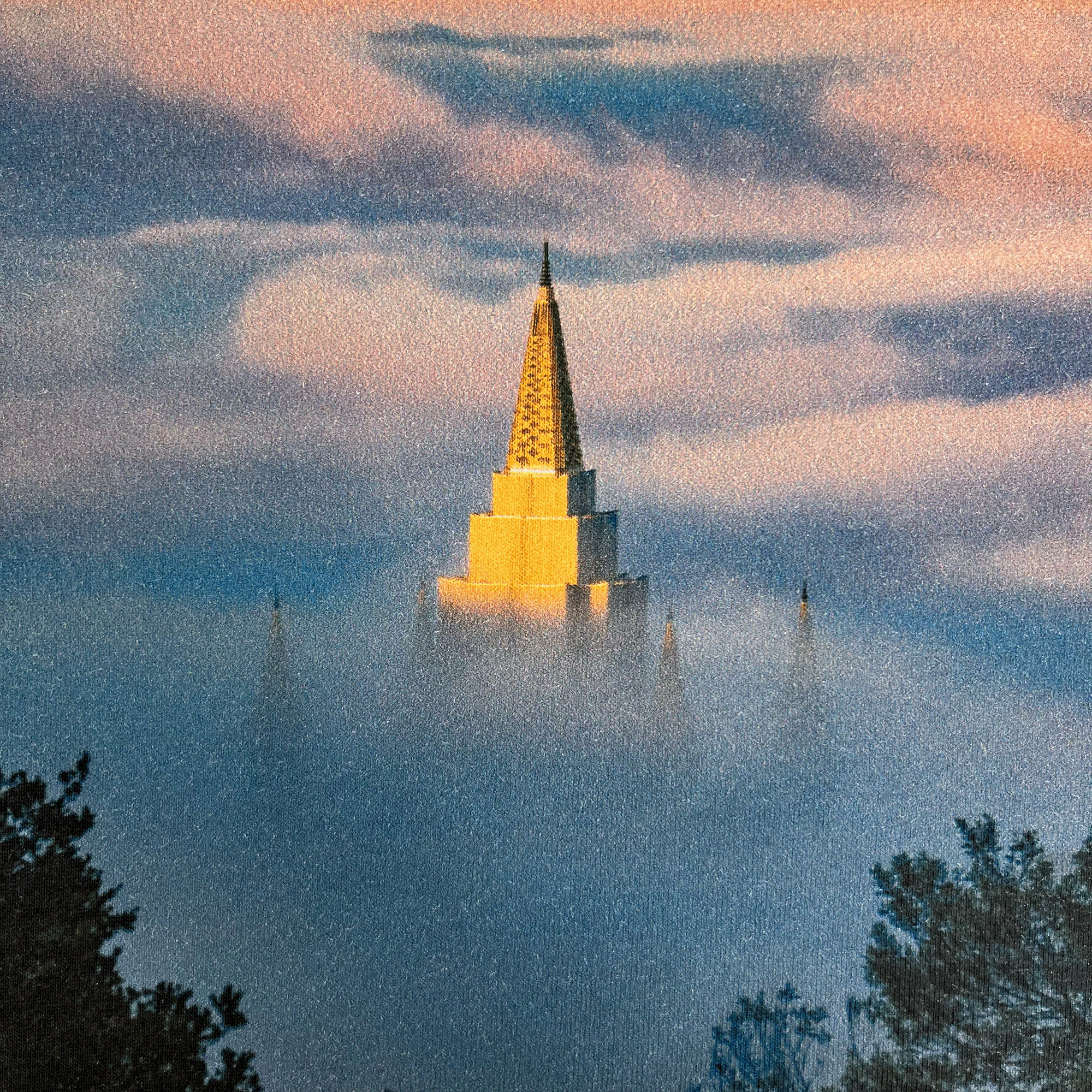 Wearing the Temple Swirl Tee by Oaklandish, you can carry with you the majestic image of a golden LDS temple spire piercing through a sea of clouds at sunrise or sunset in the Oakland Hills. The serene scene is captured perfectly, with the top of the spire softly illuminated and its lower structure enveloped in mist. Silhouettes of trees frame this ethereal moment, adding depth and tranquility to your look.