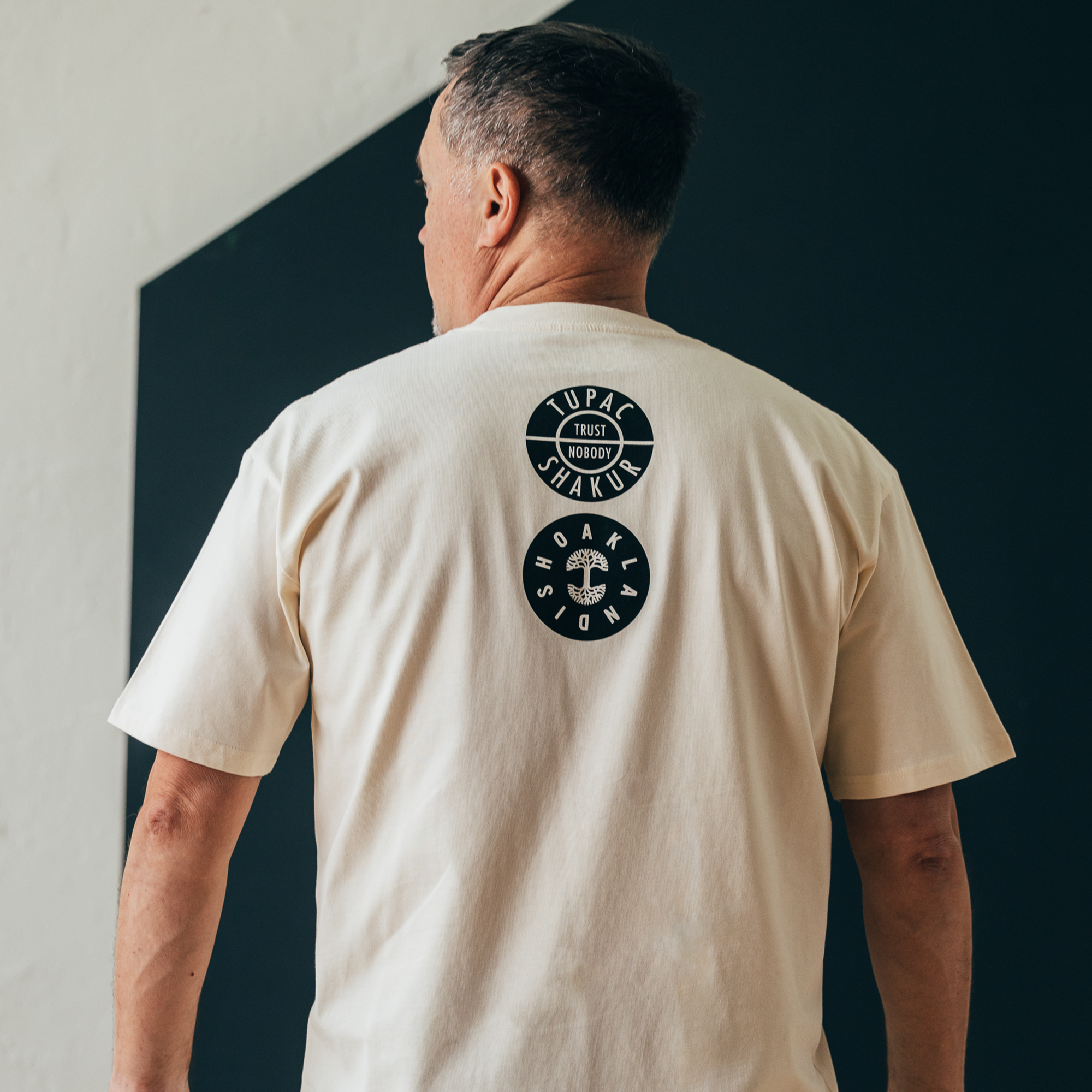 A person stands against a dark wall, wearing the Oaklandish Wavy Tupac Tee. The light-colored shirt features two circular black logos with "TUPAC SHAKUR TRUST NOBODY," incorporating small designs that celebrate hip-hop culture, showcasing only their back and part of their head as a tribute to Oakland's legacy.