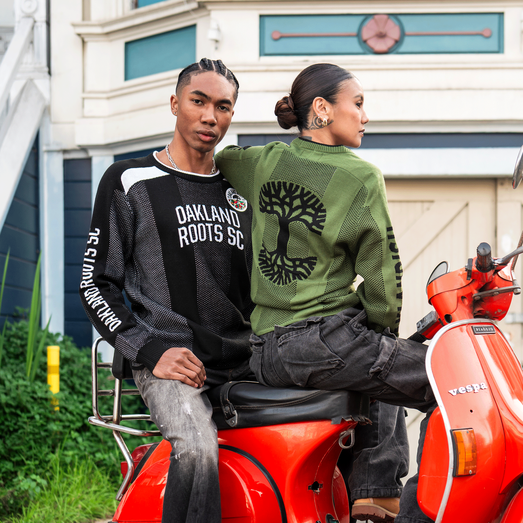 Two stylish young adults sit on a red Vespa. He wears a black Oakland Roots SC knit sweater; she sports an olive-green Oaklandish Terrace Sweater with tree design. Both are unisex, perfect for casual moments by a geometric garage door.