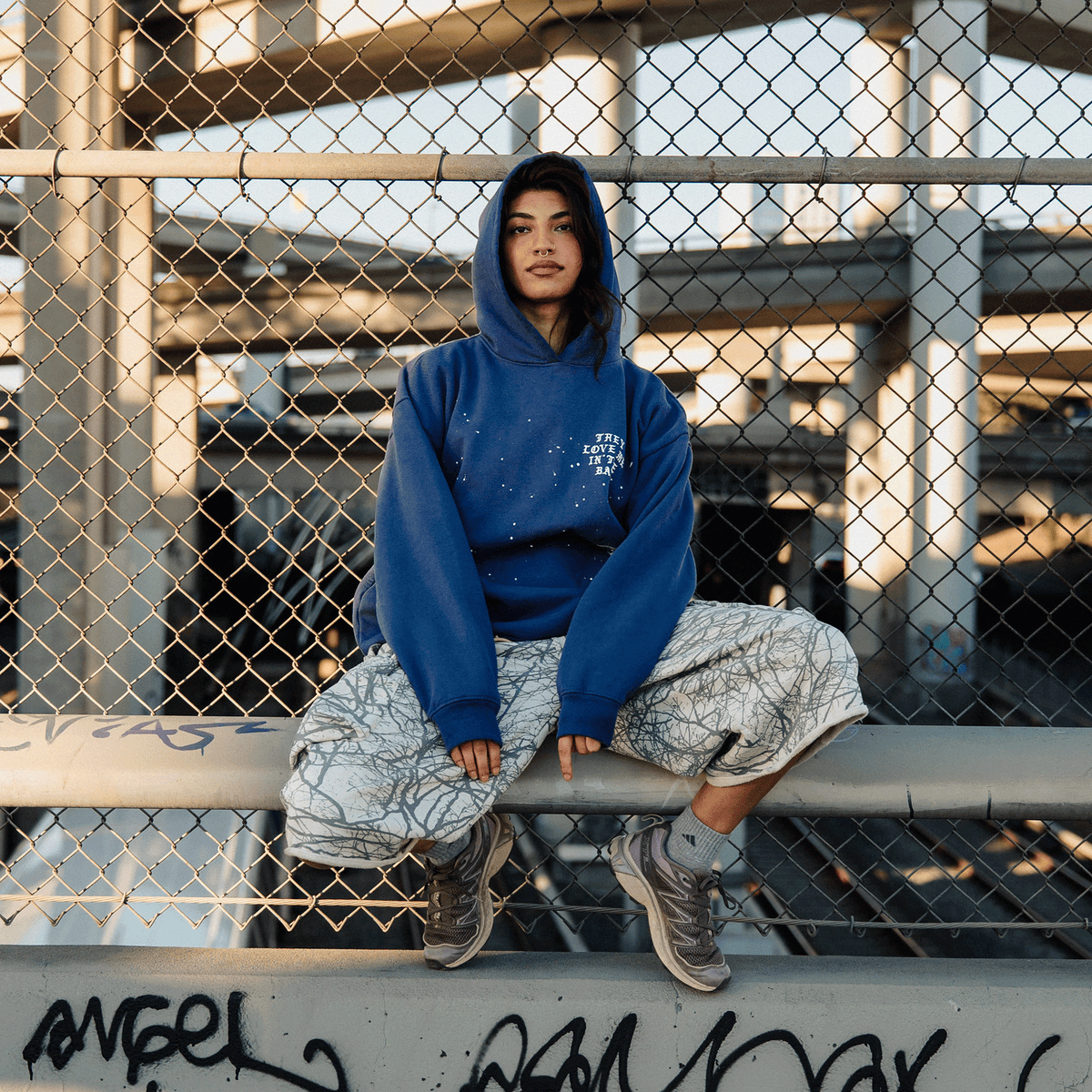 A person sits on a concrete barrier in front of a chain-link fence, wearing a vintage blue They Love Me Hoodie By Doc and patterned, loose-fitting trousers. They have dark hair, a septum piercing, and are wearing gray sneakers against an urban background of highway overpasses.