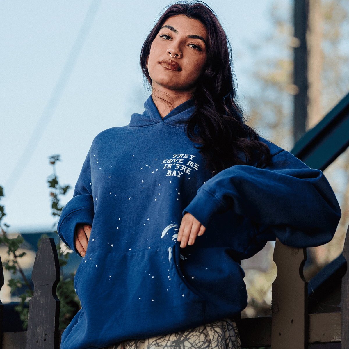 A person with long dark hair and a septum ring wears an Oaklandish They Love Me Hoodie By Doc, a vintage blue heavyweight fleece hoodie with white paint splatters and “THEY LOVE ME IN THE BAY” on the chest, leaning against a dark wooden fence with soft-focus foliage in the background.