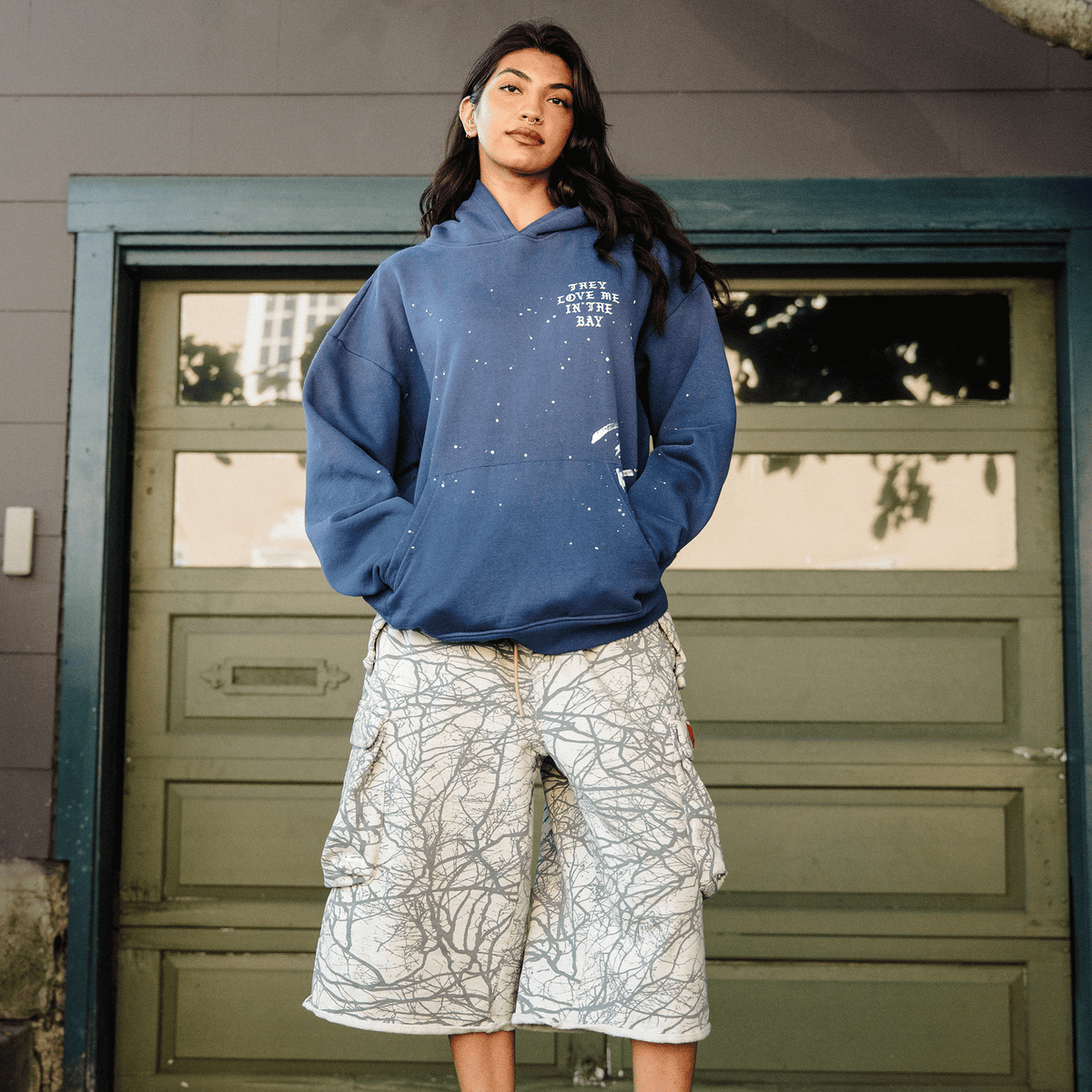 A person with long dark hair poses in an Oaklandish They Love Me Hoodie By Doc, a vintage blue heavyweight fleece pullover with white speckles. They wear loose, white knee-length shorts featuring a gray branch-like pattern, standing before a green garage door.