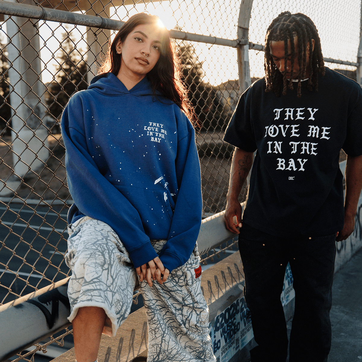 A woman in a blue hoodie with white paint splatters and text stands against a chain-link fence at sunset next to a man wearing the They Love Me Tee By Doc, a black classic fit t-shirt with bold white lettering reading, “THEY LOVE ME IN THE BAY.”