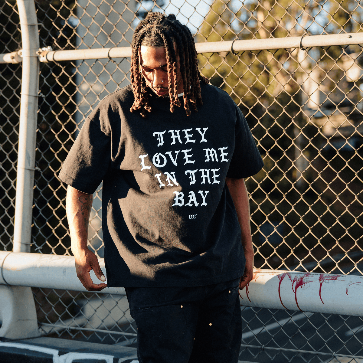 A person with long, dark dreadlocks wears a black They Love Me Tee By Doc with white text on the chest, standing outdoors in front of a chain-link fence during golden hour while wearing black pants and looking downward.
