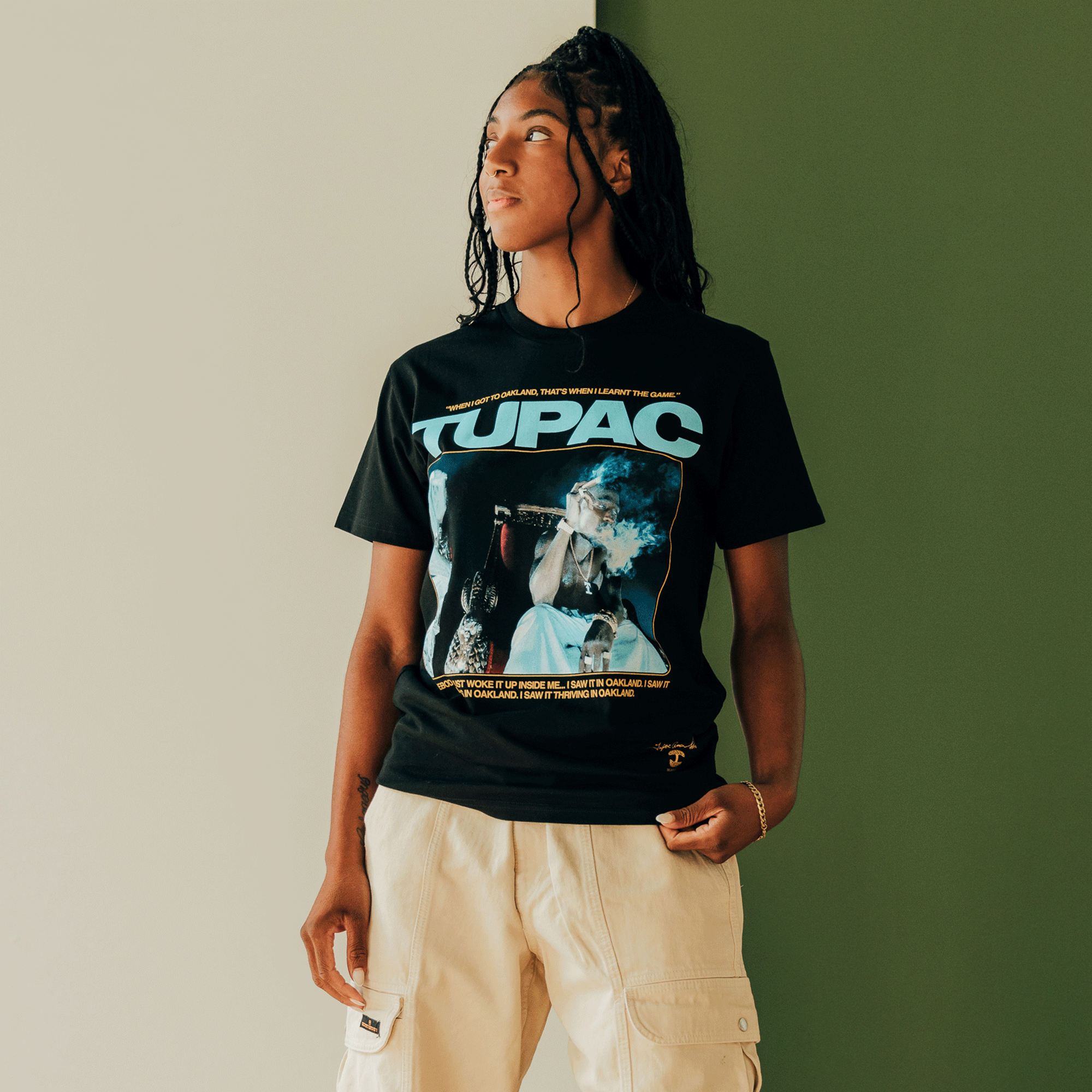 Standing against a two-tone background, a person with long braided hair exudes a hip-hop vibe. They are wearing the "Throne Tee," a black T-shirt from Oaklandish, adorned with Tupac Shakur's image and vibrant blue and orange graphics. Paired with beige cargo pants, the individual gazes to the side with a relaxed expression.