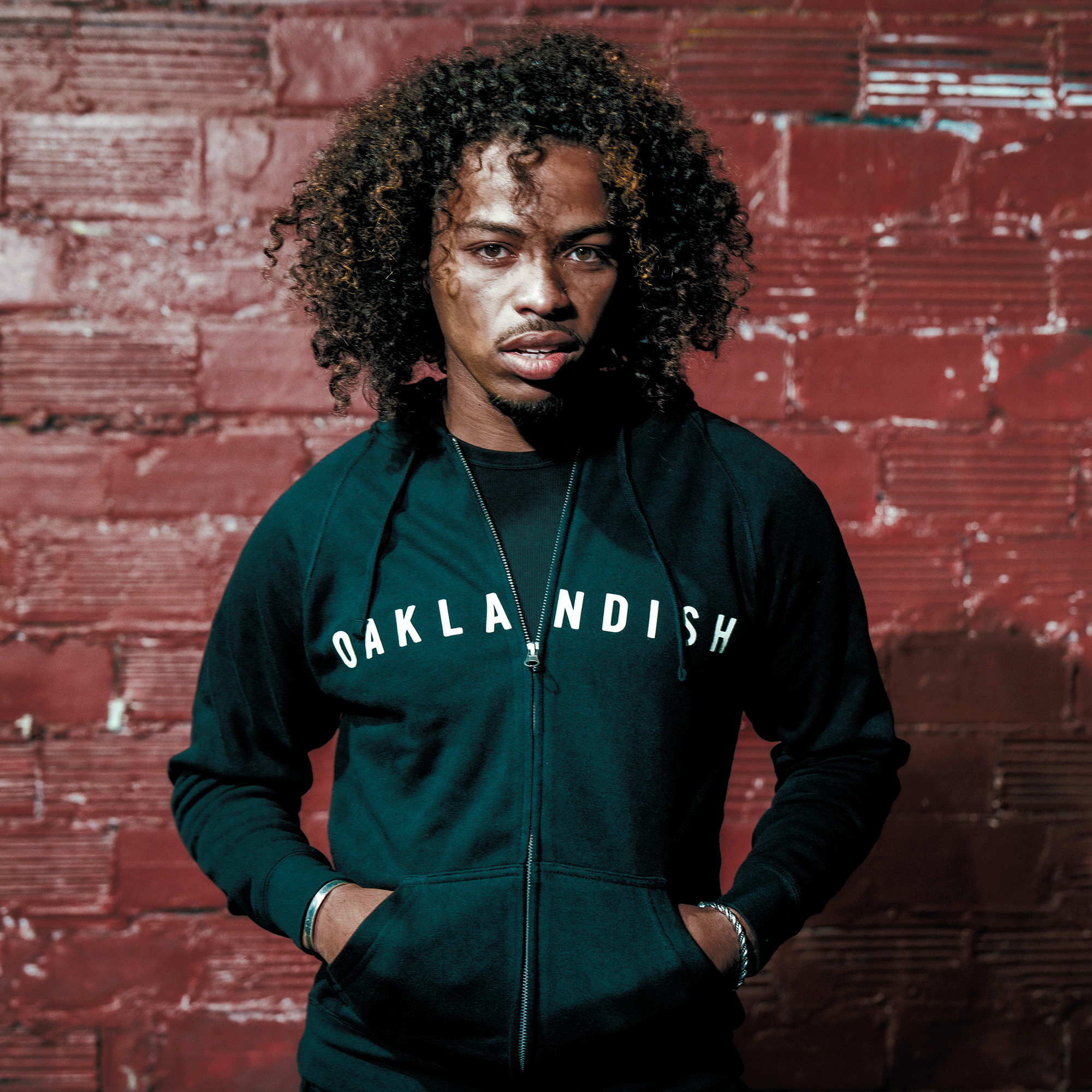 A person with curly hair wears the Oaklandish Classic Zip Hoodie by Oaklandish, standing against a red brick wall. Facing the camera with a serious look and hands in pockets, soft natural light highlights their features.