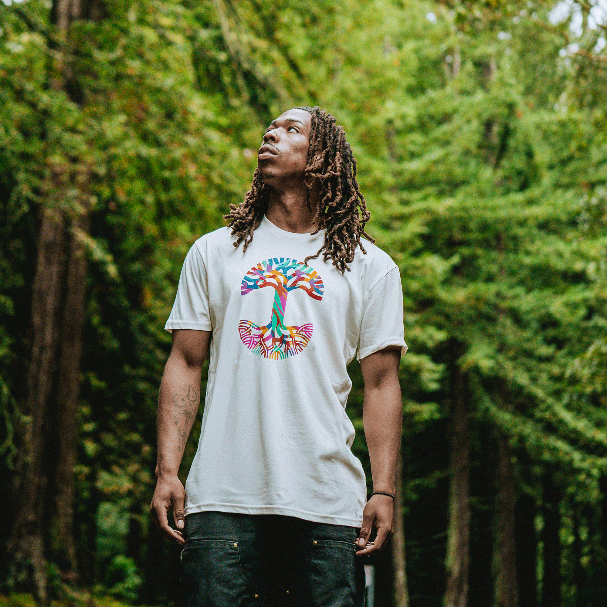 A man with long, brown dreadlocks stands in a forest, looking up. He wears the Urban Tropics Tee by Oaklandish featuring a colorful tree and roots design—a tribute to multicultural art amid lush greenery.