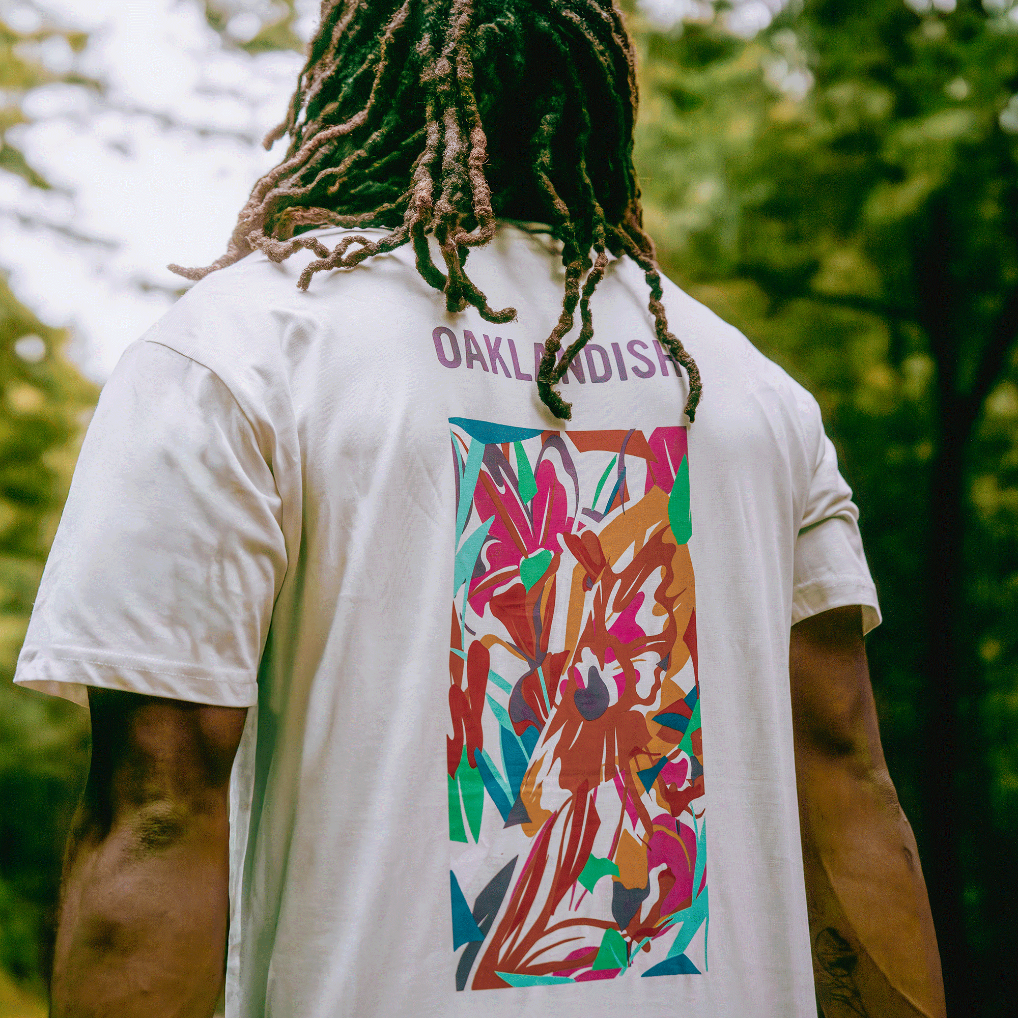 A person with long dreadlocks stands outdoors in front of green trees, wearing the Oaklandish Urban Tropics Tee with a bold, colorful design on the back.