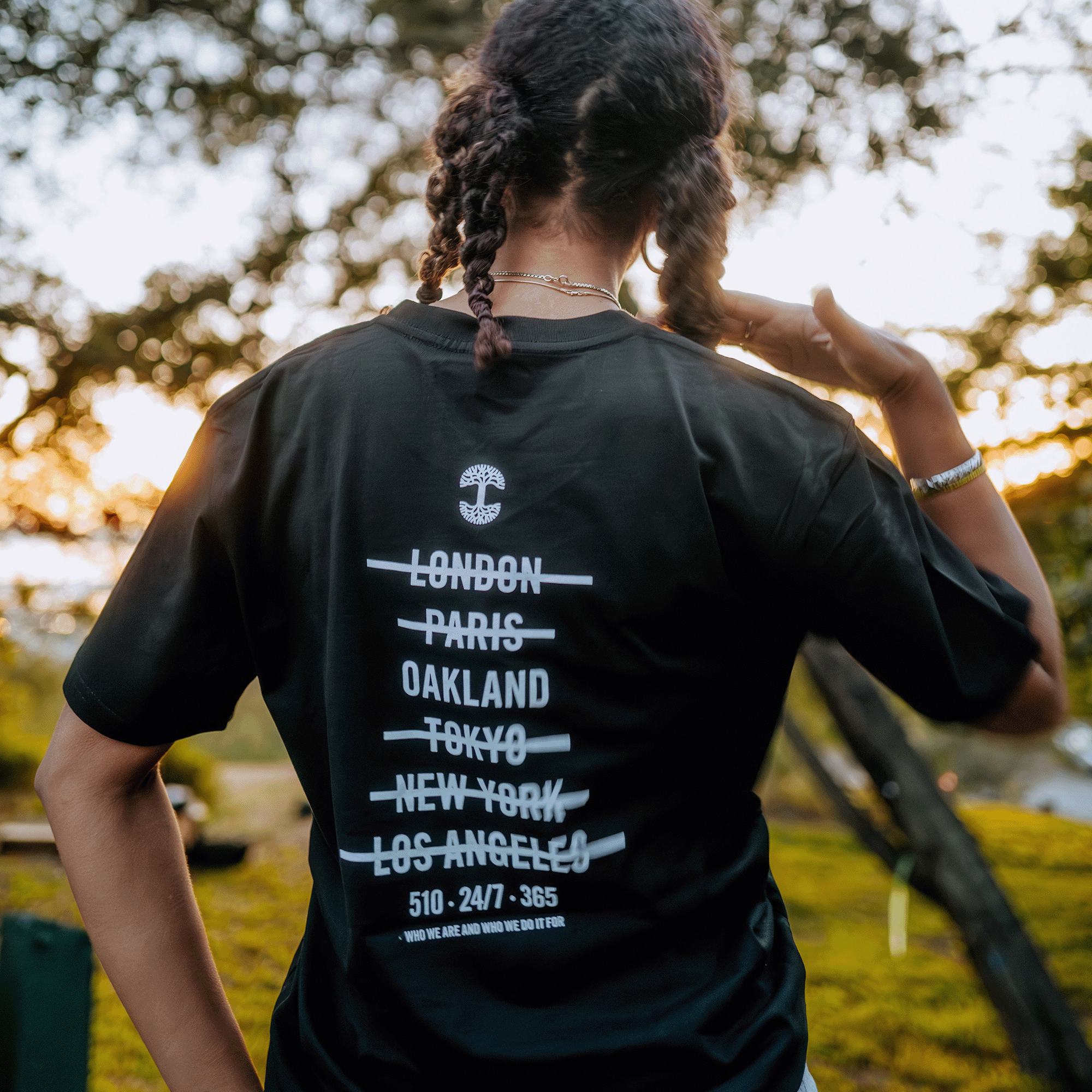 As the sun sets, a person with braided hair stands outdoors, donning an Oaklandish "Who We Are and Who We Do it For Tee" in classic black made from 100% cotton. The shirt displays crossed-out city names such as London, Paris, Oakland, Tokyo, New York, and Los Angeles. Gentle sunlight streams through the trees, adding to the peaceful ambiance.