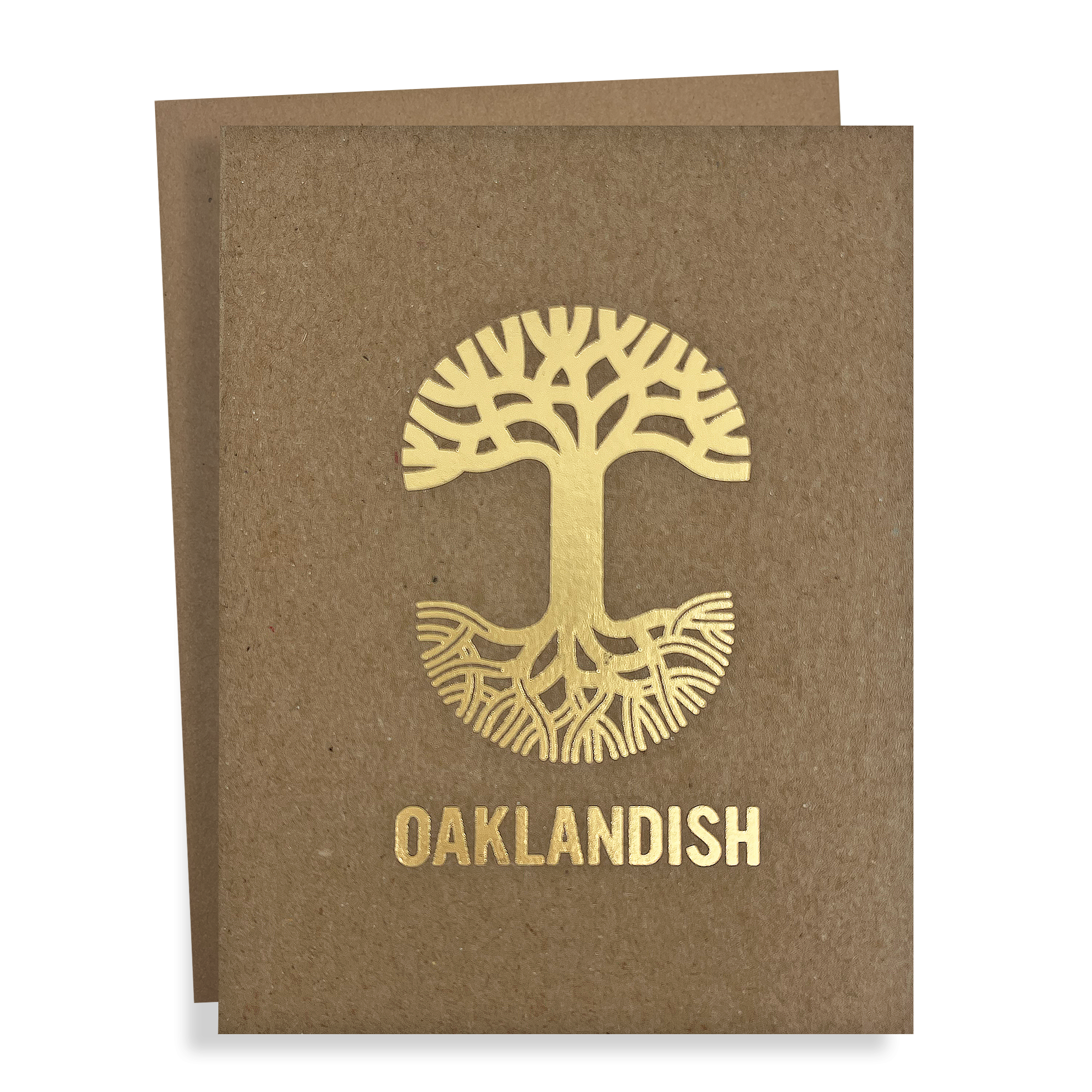 The "Oaklandish New Logo Card" by Never Elsewhere is a greeting card made from recycled brown kraft paper. It features a golden, stylized tree with roots extending in a circular design, and prominently displays the text "OAKLANDISH" below the tree. Another card slightly peeks out from behind, suggesting it's part of a set.