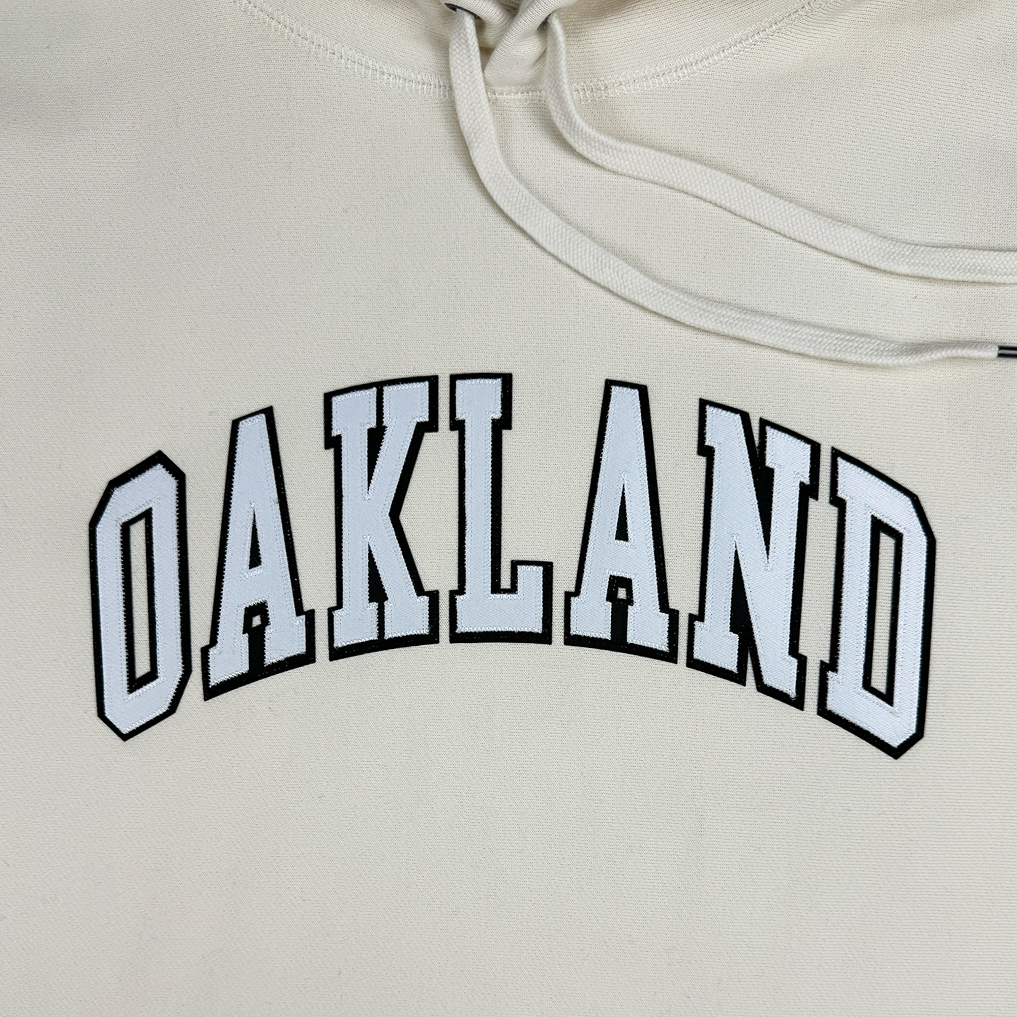 Introducing the Alma Mater Heavy Hoodie by Oaklandish: This cream-colored pullover hoodie features "OAKLAND" in bold, arched black letters outlined in white, prominently displayed on the front. The visible drawstrings enhance the hood's design, while the fleece fabric offers a soft and slightly textured feel.