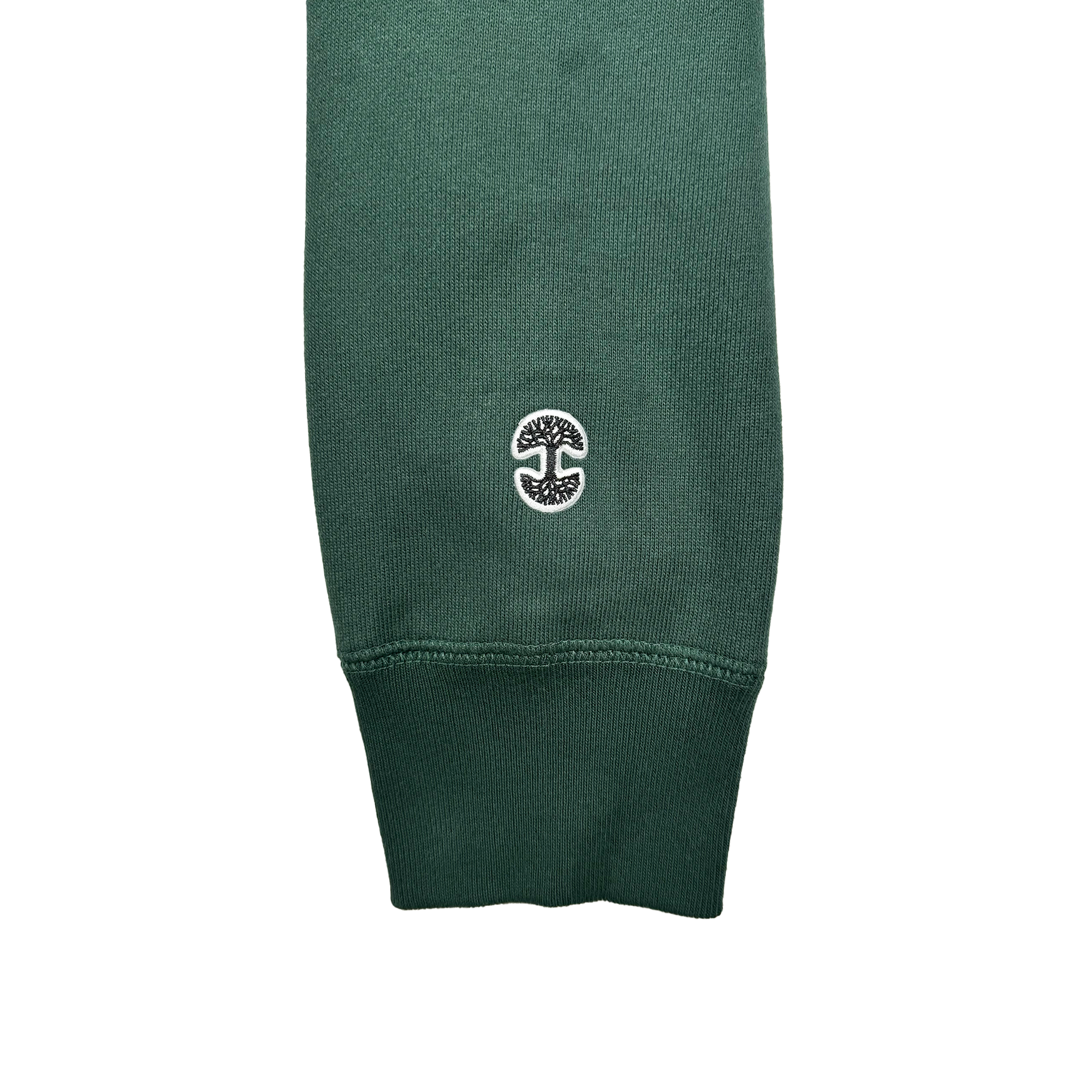 A close-up of the sleeve on an Alma Mater Heavy Hoodie, in a forest green knit, showcases a ribbed cuff. Just above the cuff lies a small embroidered white logo from Oaklandish, featuring an abstract tree design within a circular border that evokes Oakland's natural beauty. The fabric's texture displays a fine weave and subtle sheen.