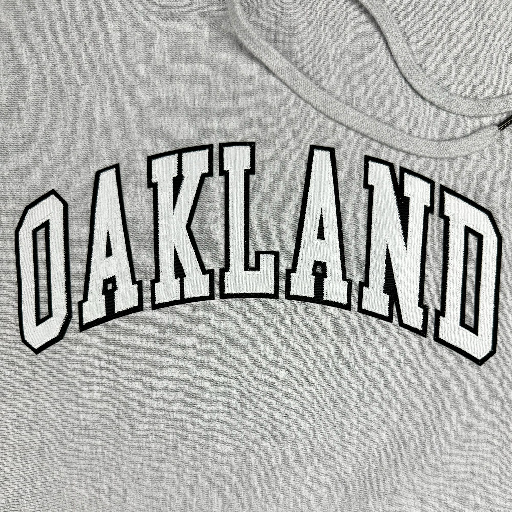 Alma Mater Heavy Hoodie by Oaklandish: A gray pullover featuring "OAKLAND" printed in bold white letters outlined in black across the front. The text elegantly arches over a soft, slightly textured fabric that suggests heavyweight fleece, complete with a drawstring for added cozy charm.