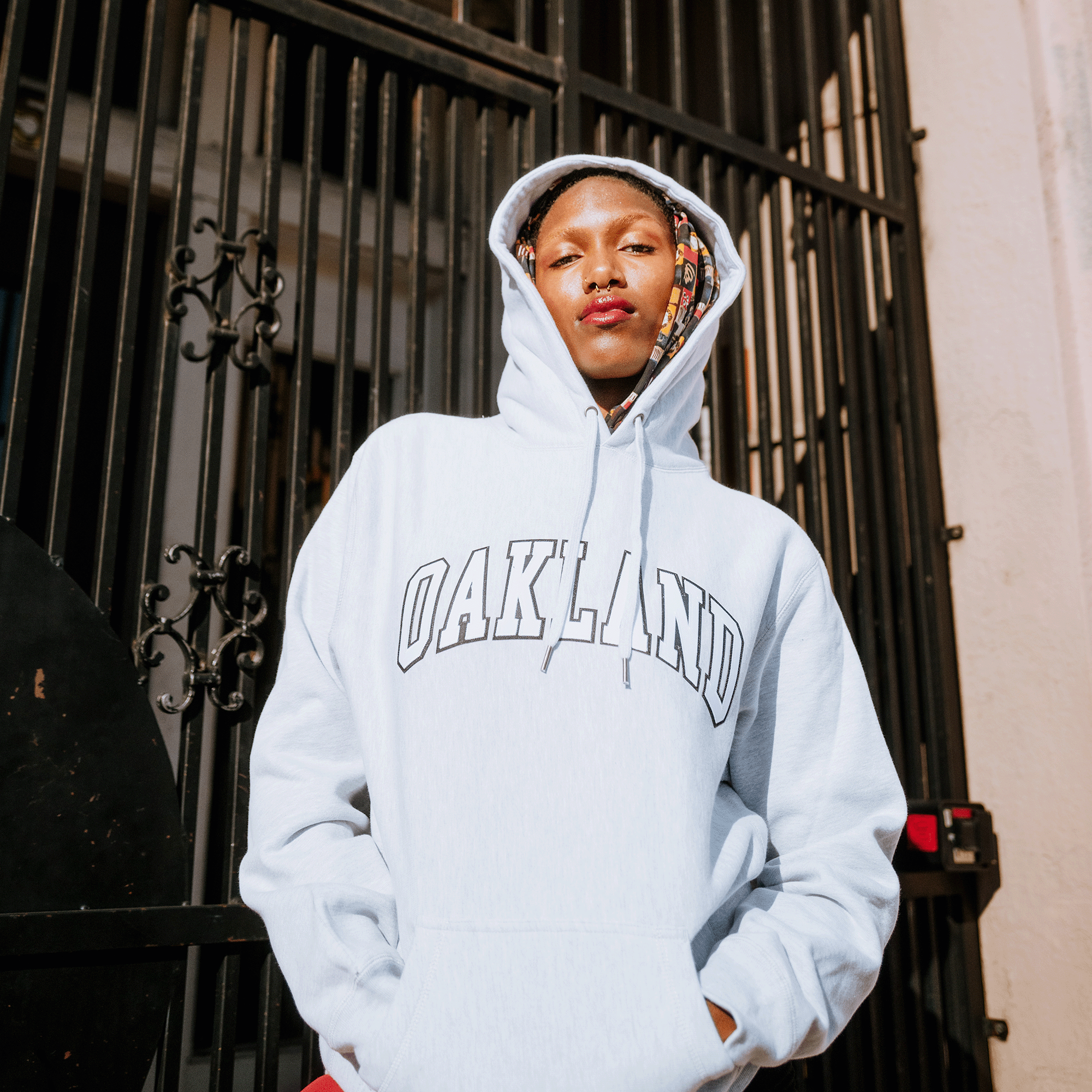 A person stands confidently in front of a metal gate, wearing a light gray Alma Mater Heavy Hoodie by Oaklandish with the hood up. A patterned scarf adds flair to the heavyweight fleece, while bright lighting highlights their face and attire perfectly.