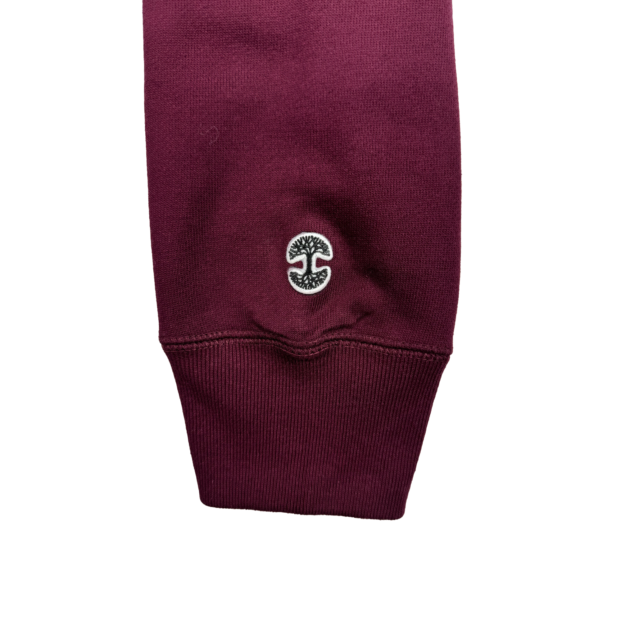 A detailed view of a sleeve from the Oaklandish Alma Mater Heavy Hoodie in maroon, featuring a ribbed cuff. Close to the cuff, there is an embroidered circular logo depicting a tree with mirrored branches and roots in black and white. The smooth fabric highlights intricate stitching against a plain white background.