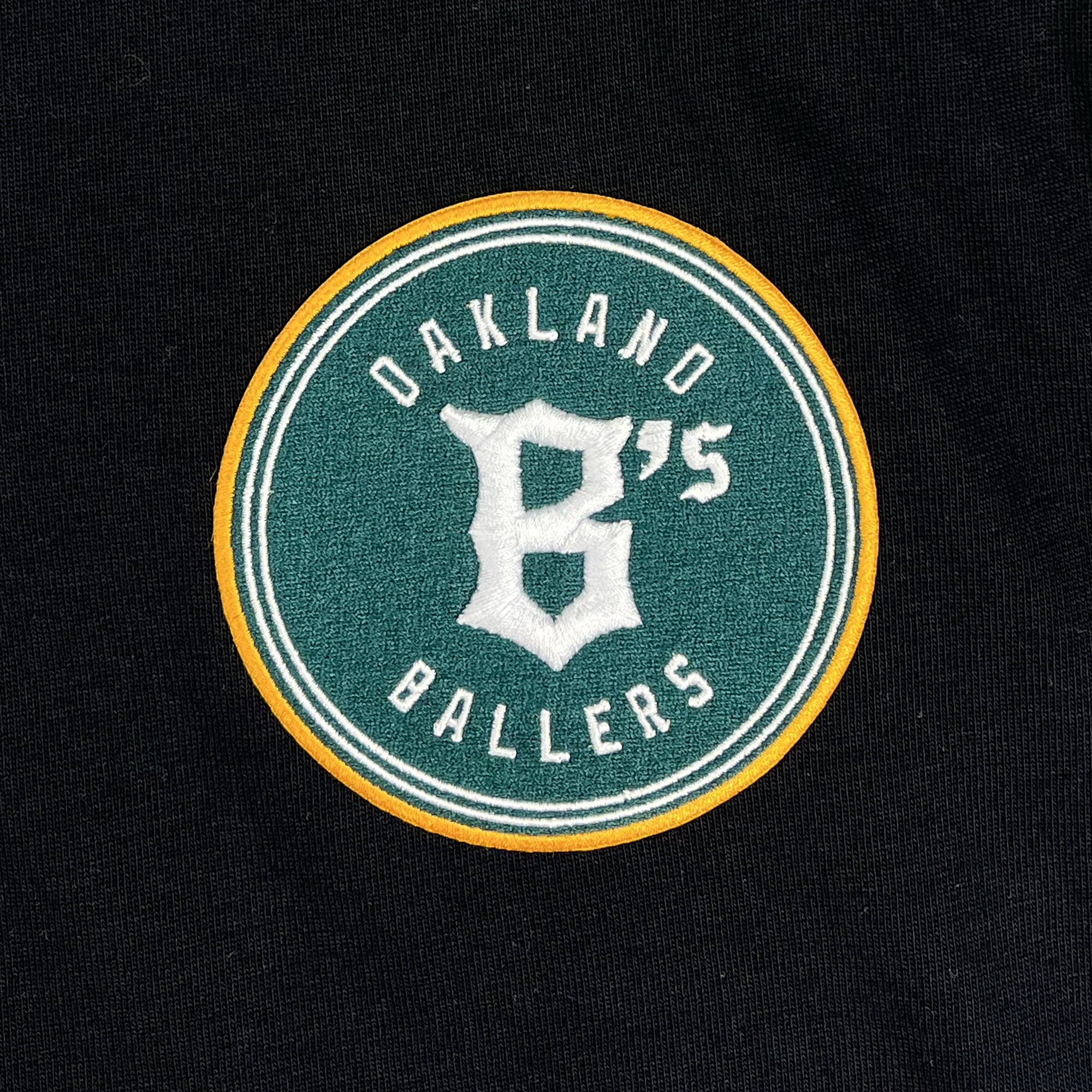 A green circular patch with a yellow border is stitched onto the black fabric of the Oakland Ballers Heavy Baseball Jersey. The text "OAKLAND BALLERS," representing the professional baseball team in the Pioneer League, appears in white around the perimeter with a large white "B" followed by an apostrophe and the number "5" at its center.