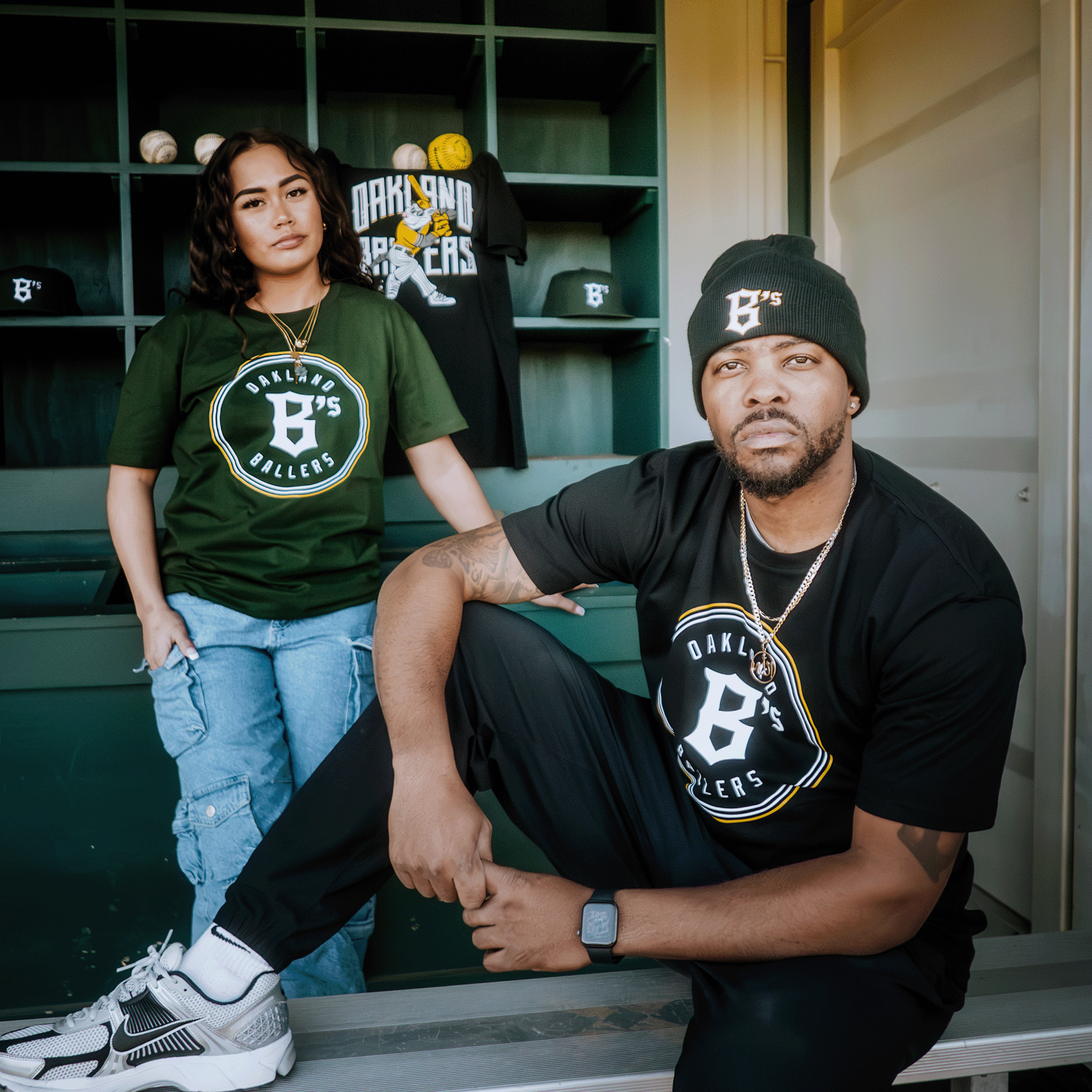 A man and woman in casual attire are in front of a dugout. The man, wearing a beanie and an Oakland Ballers Classic Tee, sits with one knee up. The woman stands in the same tee and ripped jeans. Shelves behind them display sports gear illustrating their love for the Oakland Ballers team.