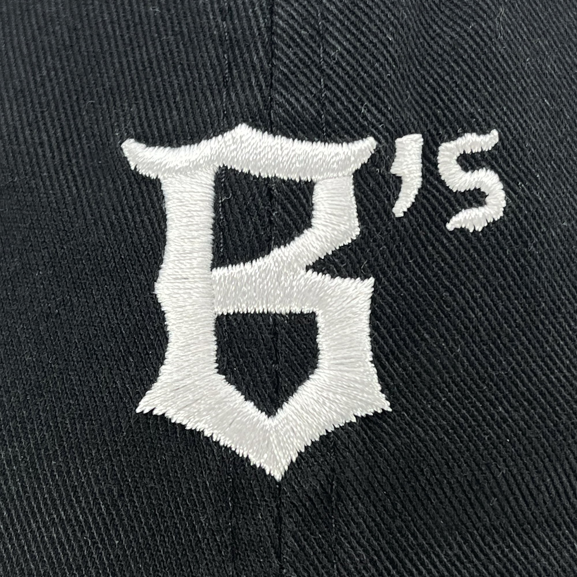 Close-up of the Oakland Ballers Dad Hat, showcasing a black fabric with an embroidered white logo. The embroidery features a Gothic-style letter "B" with an apostrophe and the letter "S" to the upper right of it, representing the Oakland Ballers, a new professional baseball team in the Pioneer League. The dense stitching contrasts sharply with the black background.