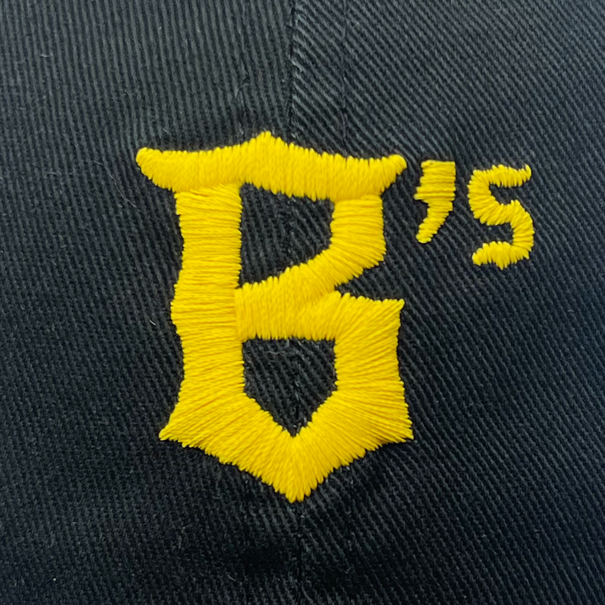 A close-up image of the Oakland Ballers Dad Hat, showcasing its black fabric with an embroidered yellow "B" and an apostrophe followed by an "s" in the top right corner. The bold and prominent stitching features the letter "B" in a gothic font style, reminiscent of the professional baseball team Oakland Ballers.