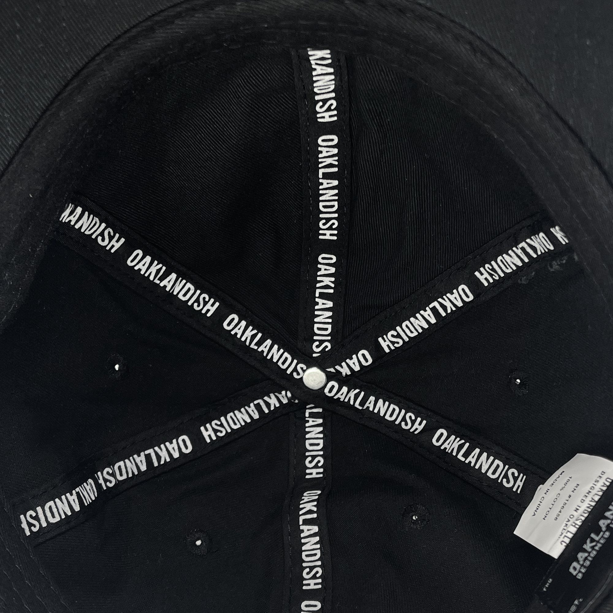 The interior of the Oakland Ballers Dad Hat features white text on the seams that reads "OAKLANDISH" repeatedly. A black button is located at the center where the seams intersect. A white tag with information celebrating the new professional baseball team, Oakland Ballers, is visible on the right side of the image.