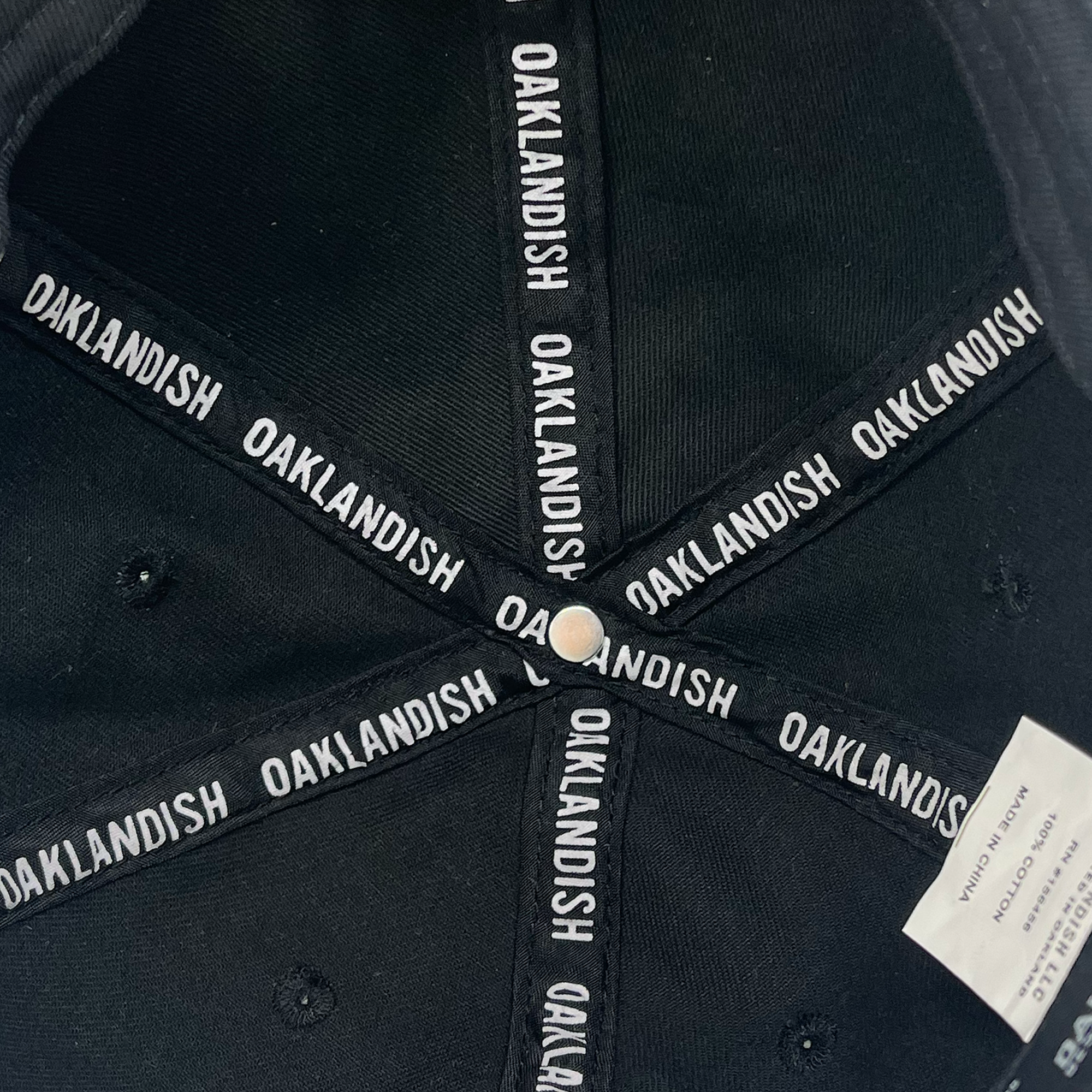 Interior view of the Oakland Ballers Dad Hat featuring white "OAKLANDISH" text repeatedly printed on the panels. At the center is a button fastener. There's an attached white tag with manufacturing details and care instructions visible on the side. The cap's stitching and construction details, reminiscent of gear worn by professional baseball teams, are also visible.