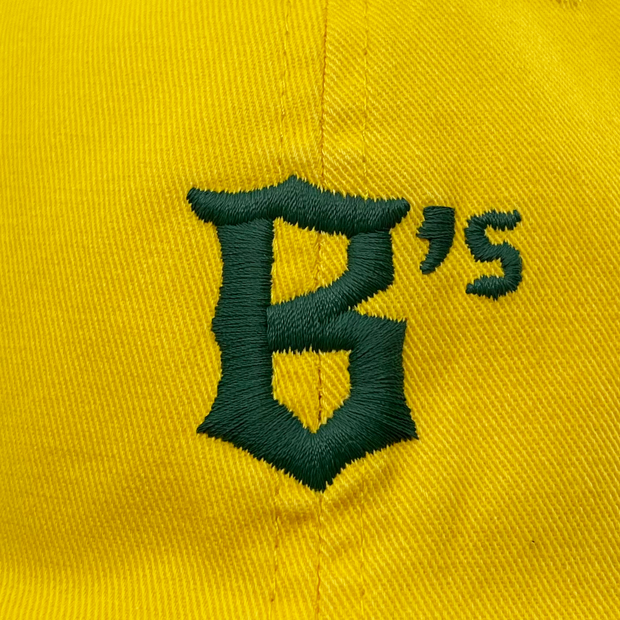 A close-up image of a yellow Oakland Ballers Dad Hat with a green embroidered "B's" logo on it. The logo, belonging to the Oakland Ballers brand, features the letter "B" in a stylized, bold font, followed by an apostrophe and the letter "s" in matching green thread. The stitches of the embroidery are clearly visible.