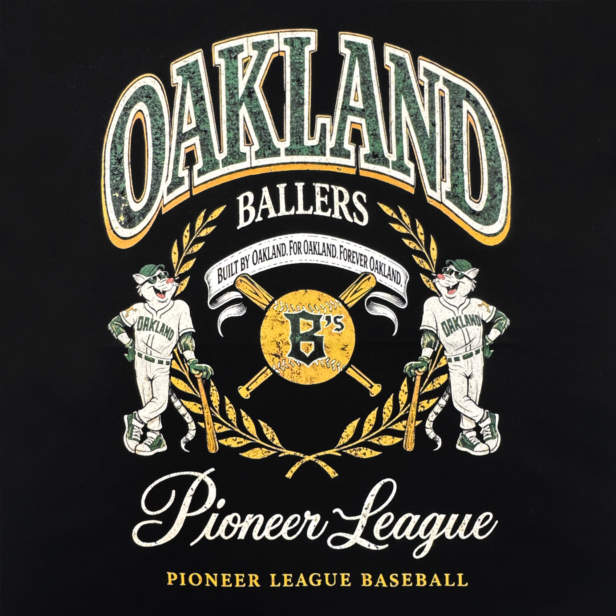 A graphic on the Oakland Ballers Double Tee features two smiling baseball mascots in Oakland uniforms, a golden bat-and-ball crest, and green/yellow accents, with "Oakland Ballers, Pioneer League Baseball" text celebrating the team.