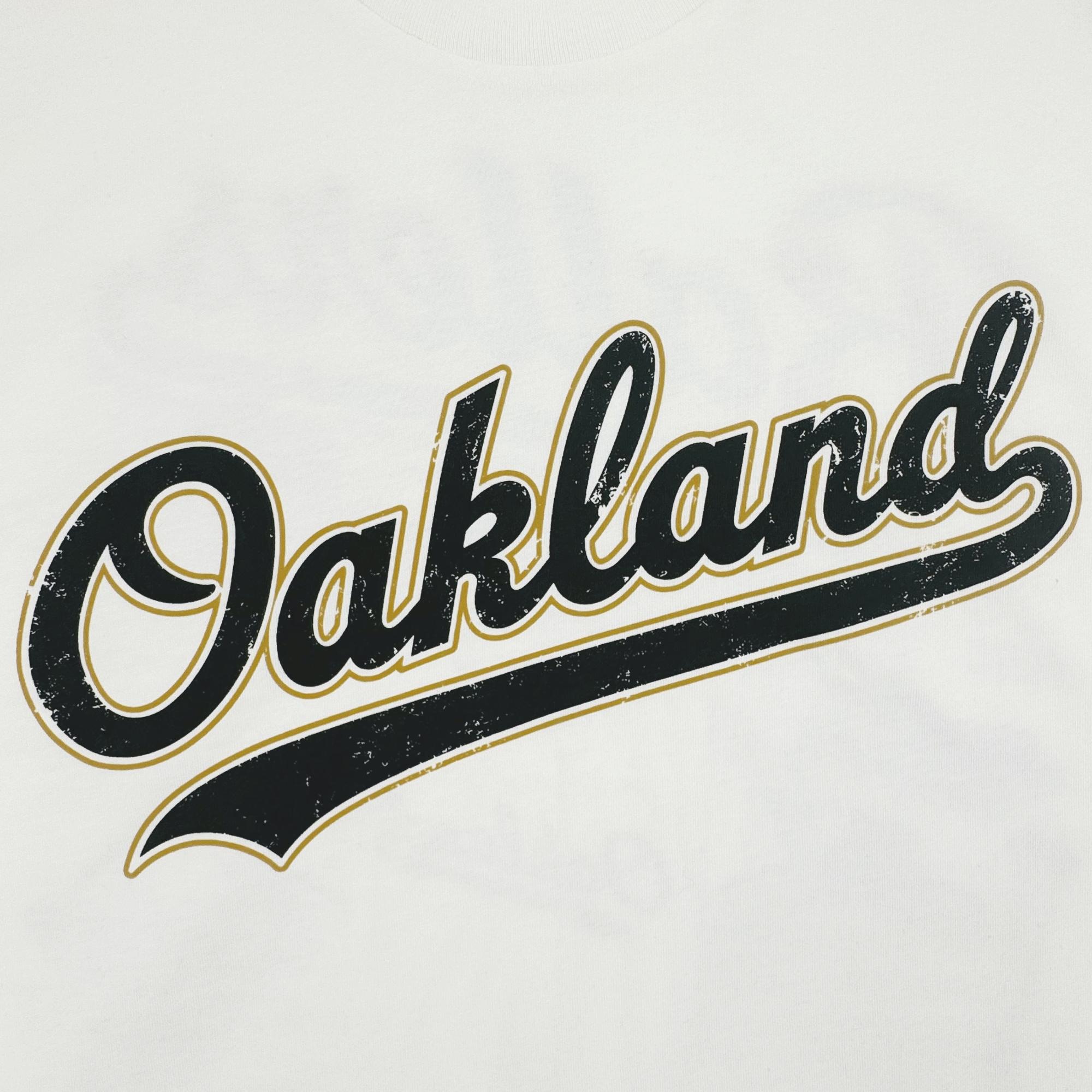 The Dugout Tee by Oakland Ballers features “Oakland” in bold black cursive with a gold outline and distressed finish on white, inspired by classic Oakland baseball team logos.