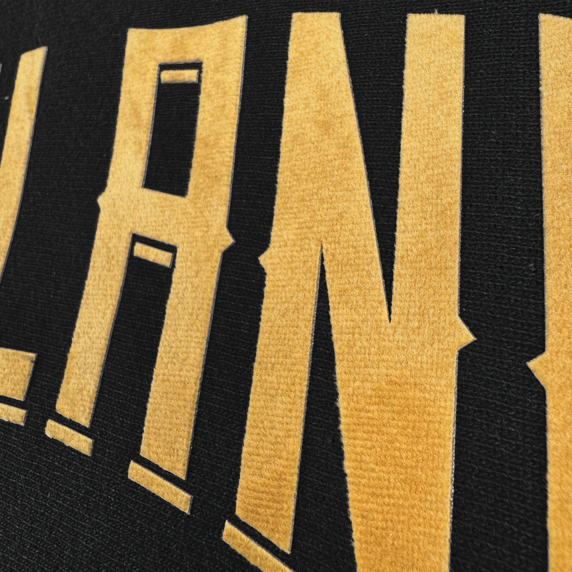 A close-up of bold, textured yellow letters on black fabric highlights Oakland Ballers' branding, featuring the Champion X Oakland Ballers Wordmark Hoodie. The letters "LAN" reveal intricate stitching and a striking contrast between dark fabric and bright lettering.