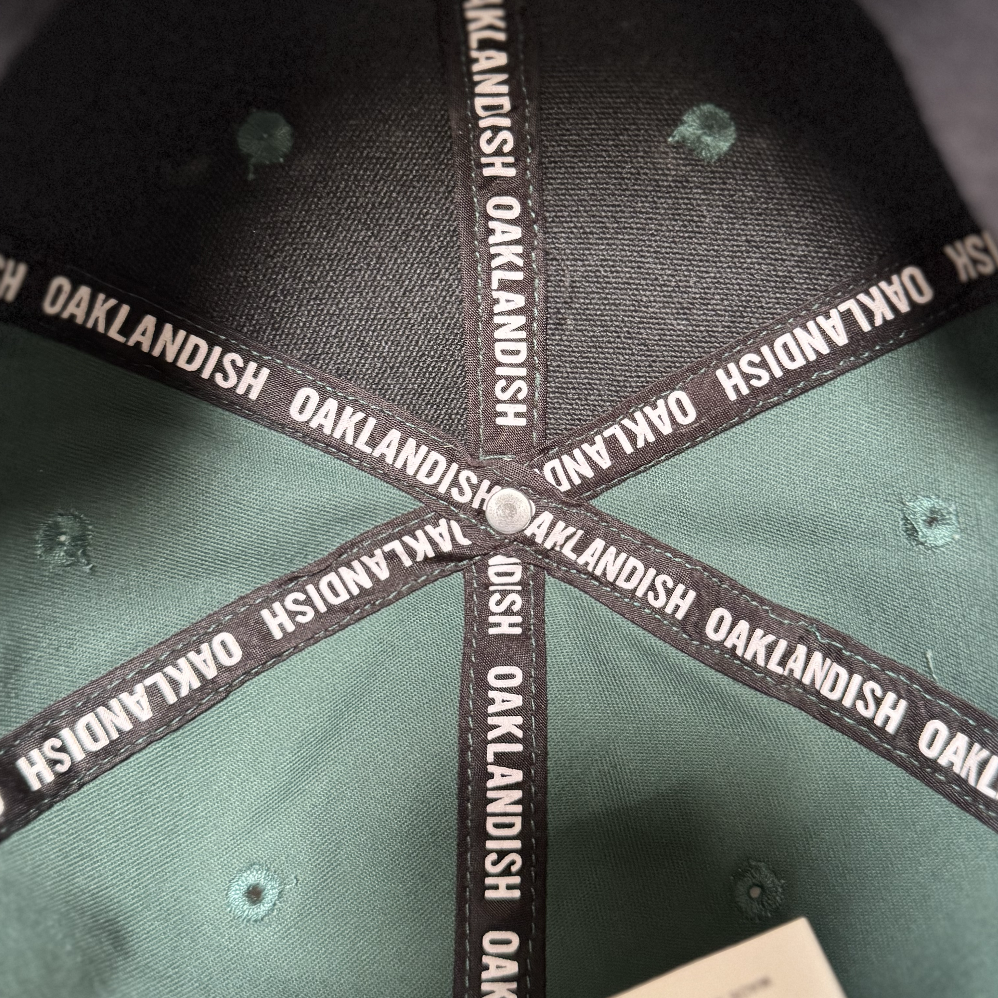Close-up of the Oakland Ballers Logo Hat’s interior reveals black fabric strips with "OAKLANDISH" in white, celebrating the Oakland Ballers baseball team. The green and black panels meet at the top, secured by a metal rivet.