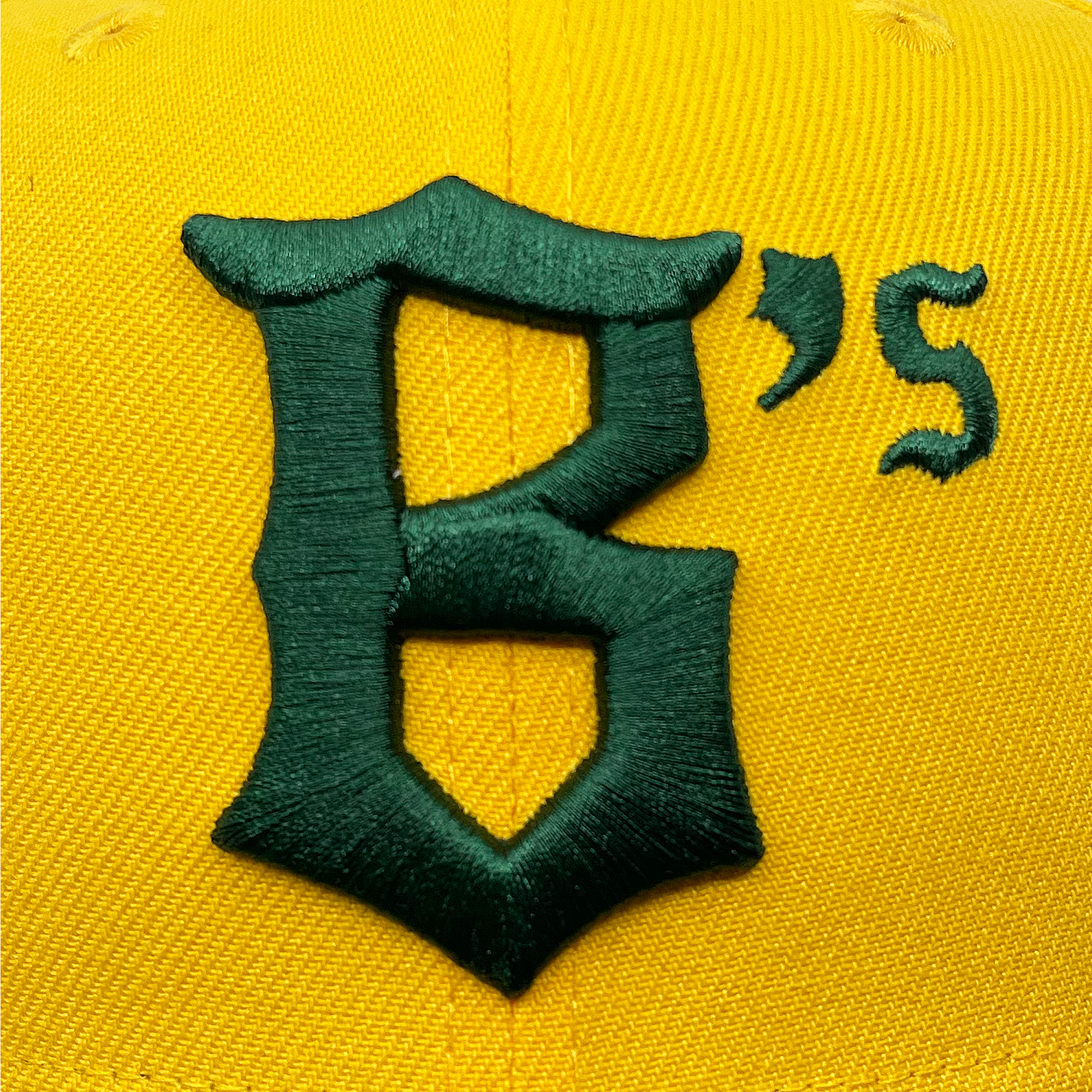 A close-up of the New Era Oakland Ballers 9FIFTY Cap shows a stylized green embroidered letter "B" followed by a small "’s" on the right. The hat's yellow fabric texture is noticeable, with clear stitching on the design. The embroidery, representing the Oakland Ballers professional baseball team, stands out against the bright yellow background.