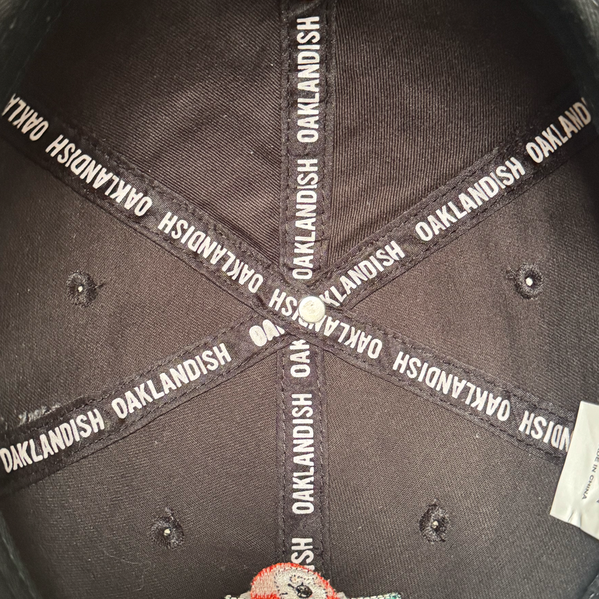 Close-up of the inside top of the black AC Transit Dad Hat by Oaklandish. Black fabric strips radiate outward, each with "OAKLANDISH" in white letters, celebrating East Bay pride. A white stitched label appears on the lower right side.