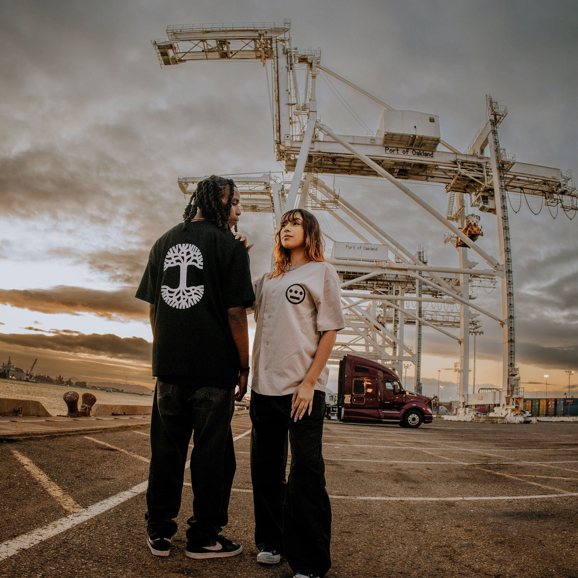 Two people stand near large industrial cranes, with a port visible in the background. One wears a black T-shirt with a circular graphic design on the back; the other sports an Oaklandish Heavy Button-up Baseball Jersey featuring the Oaklandish tree logo. Both are wearing black pants. The sky is overcast, creating a moody, twilight ambiance.
