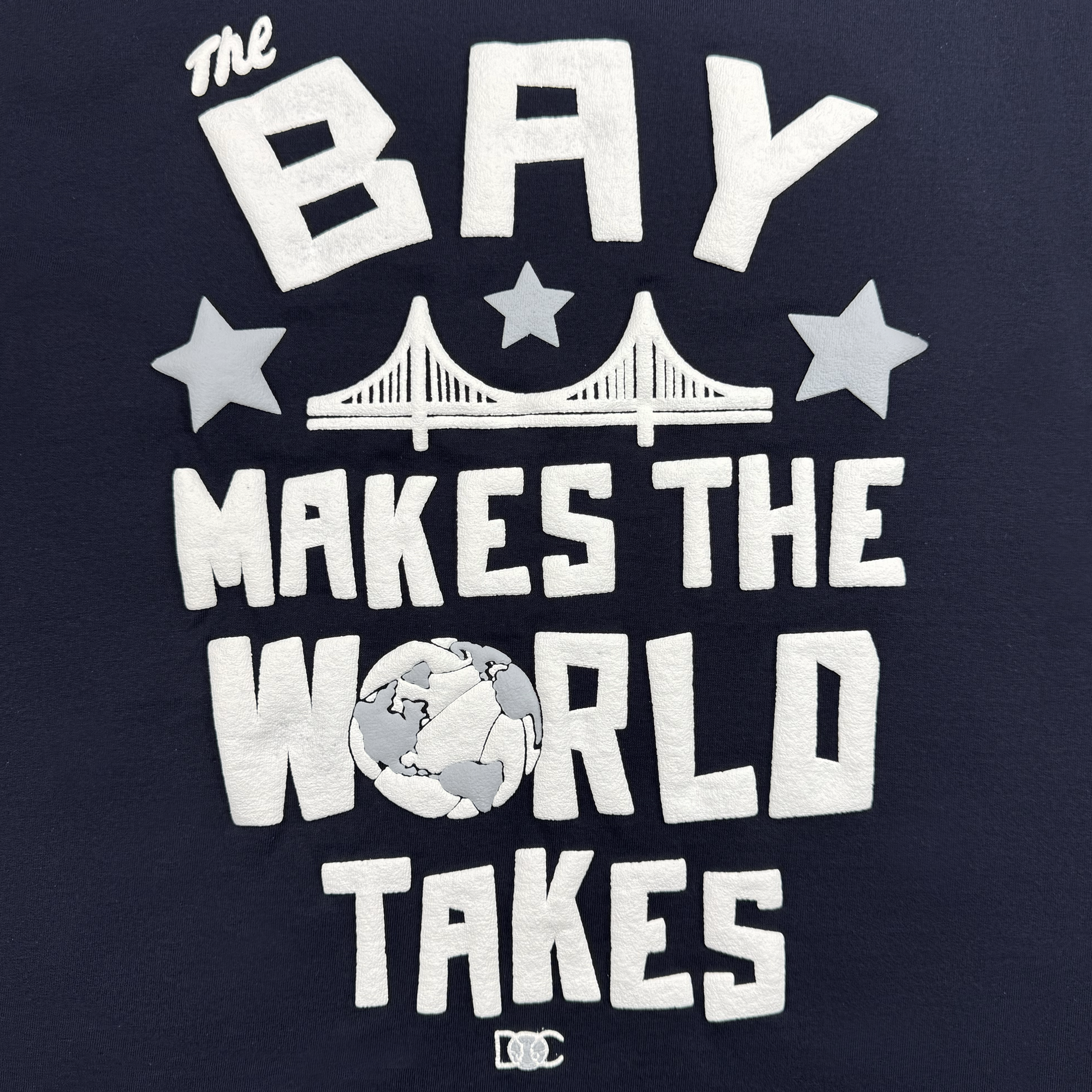 The Bay Bridge Tee by DOC from Oaklandish showcases a Bay Area-inspired design. It features "The Bay Makes The World Takes" in bold white on a dark background, with a stylized bridge and stars above, and a globe as the 'O' in "World," plus two small bridge icons at the bottom.