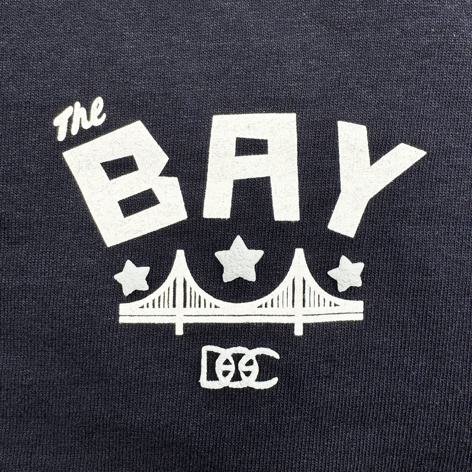 The Bay Bridge Tee by DOC from Oaklandish features a dark fabric with "The" in cursive, "BAY" in bold capitals, two stars over a stylized Bay Area bridge, and interlocked "DC" letters with a small "C," celebrating global culture through art and design.