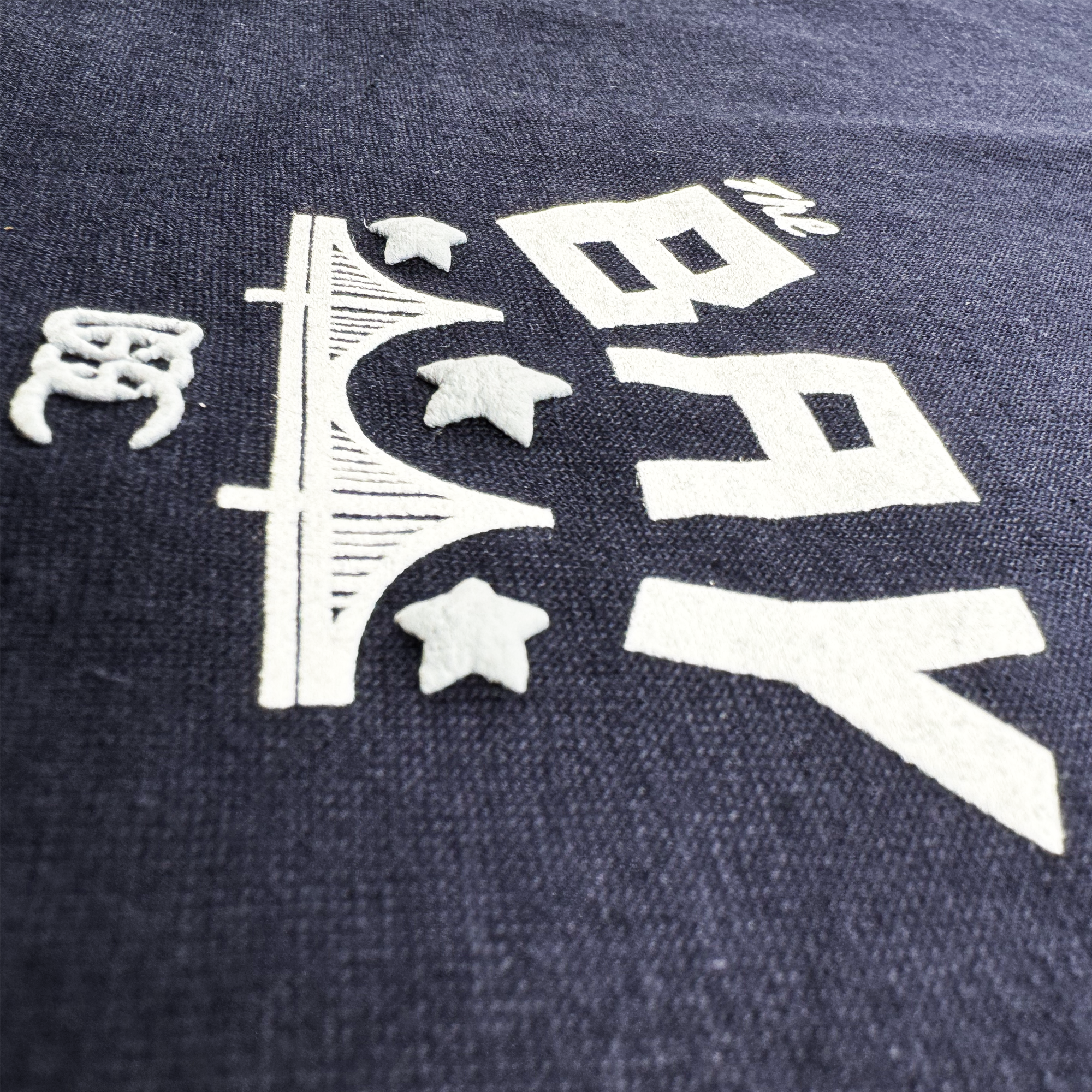 A close-up of the navy fabric of the Bay Bridge Tee by Oaklandish shows a white graphic with "THE BAY" in bold letters, featuring a stylized bridge and three stars, capturing the vibrant art and culture of the Bay Area.