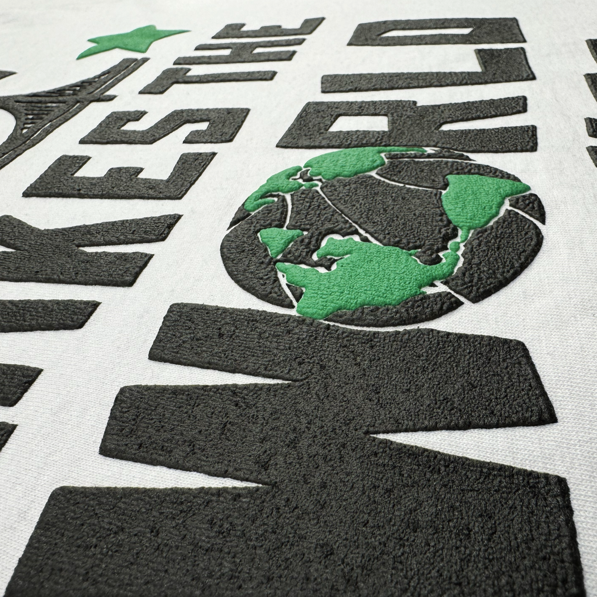 Close-up of the Bay Bridge Tee by DOC, designed by Oaklandish, showcases embroidered text "THE WORLD" with a stylized Eiffel Tower. A globe illustration with green continents and black oceans is featured near the "O," exhibiting global culture's essence in Dustin O. Canalin's signature raised style.