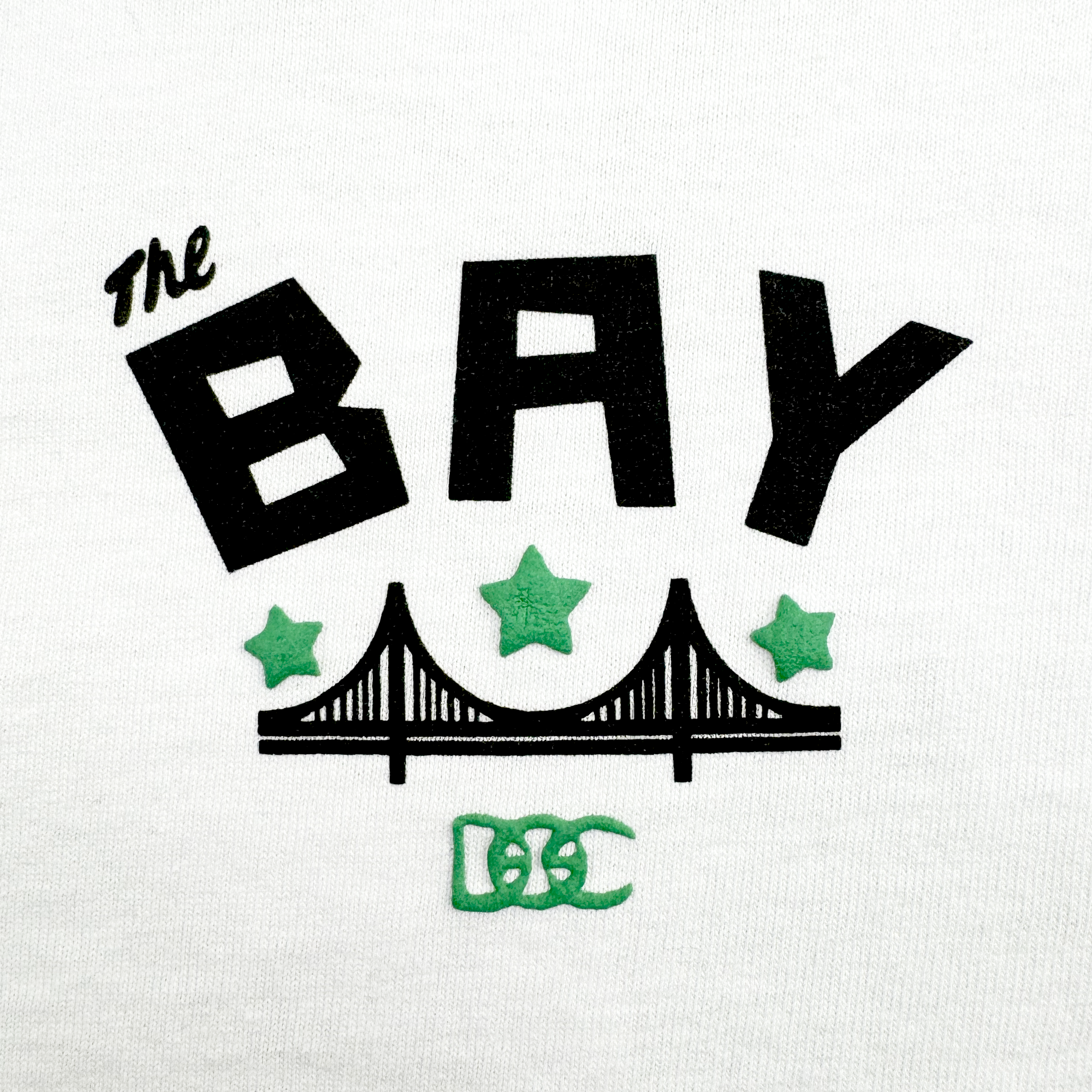 The Bay Bridge Tee by DOC from Oaklandish features bold black "The BAY" text above a bridge illustration with Bay Area vibes, three green stars, and two interlocking green C's at the bottom on a white background.