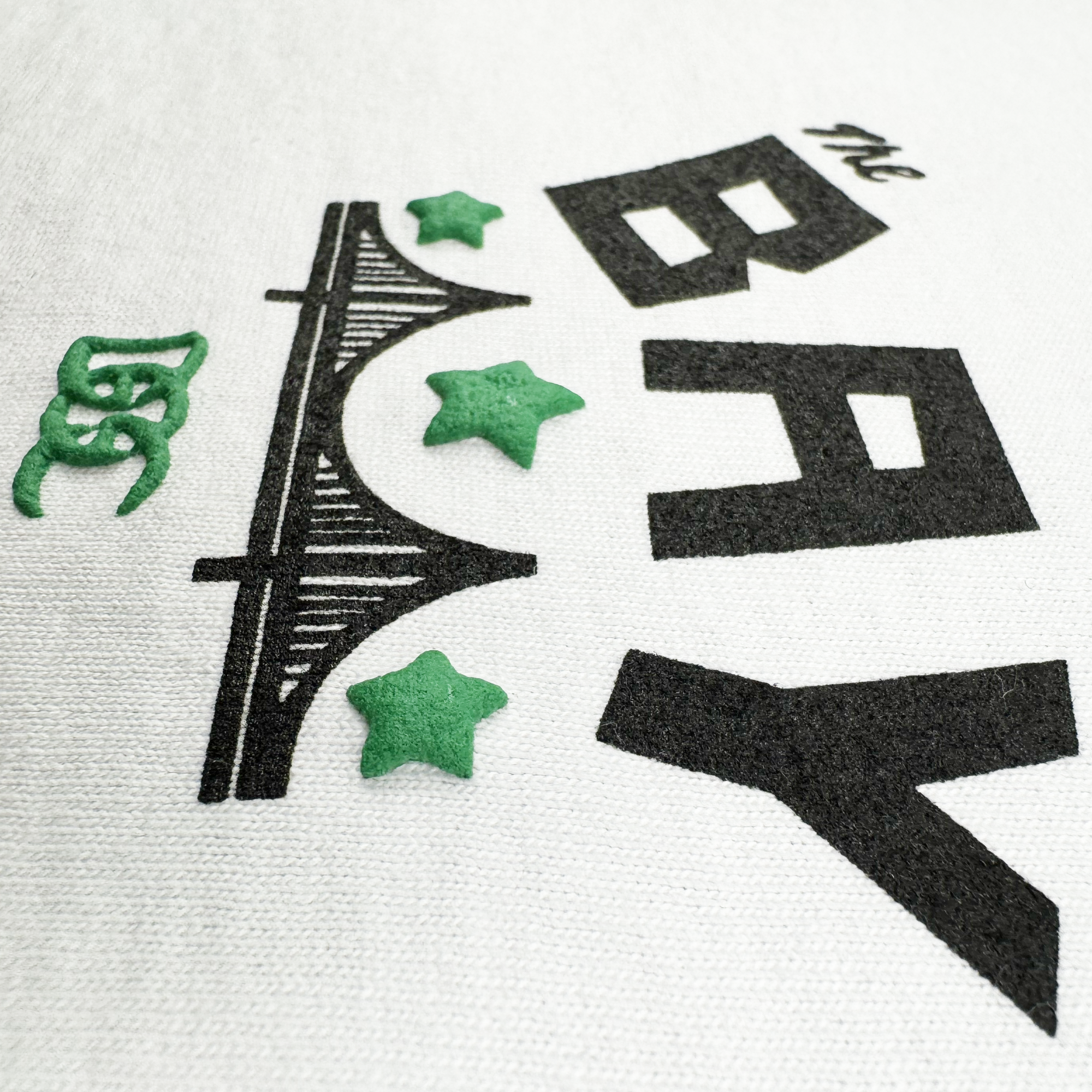 The Bay Bridge Tee by DOC from Oaklandish features a design with "The Bay" in bold black on white fabric, a stylized black bridge underneath, and accents of three green stars and a small emblem. This homage to Bay Area culture is envisioned by Dustin O. Canalin.