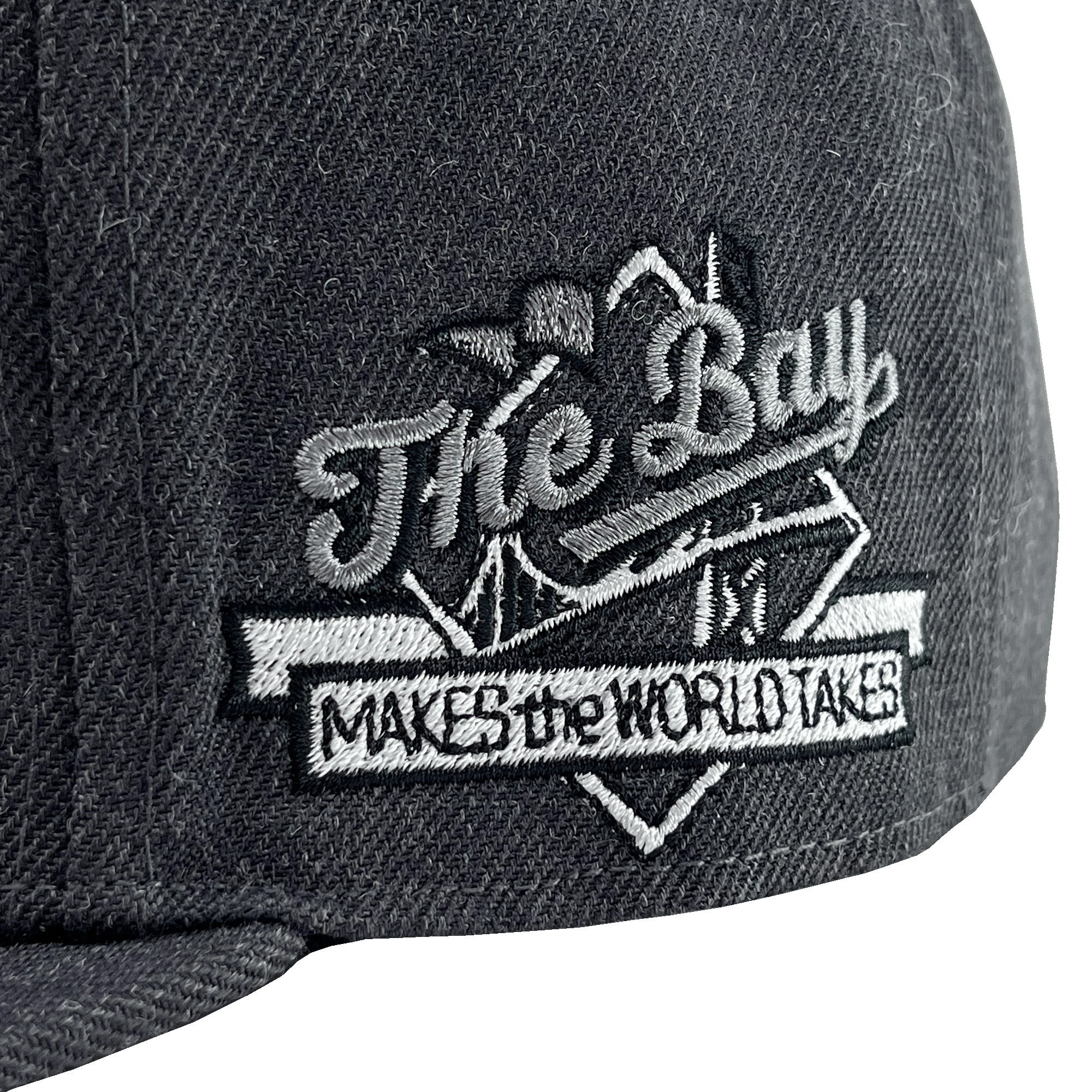 Close-up of The Bay Makes, The World Takes detail on the wearer’s left side of a charcoal hat. 
