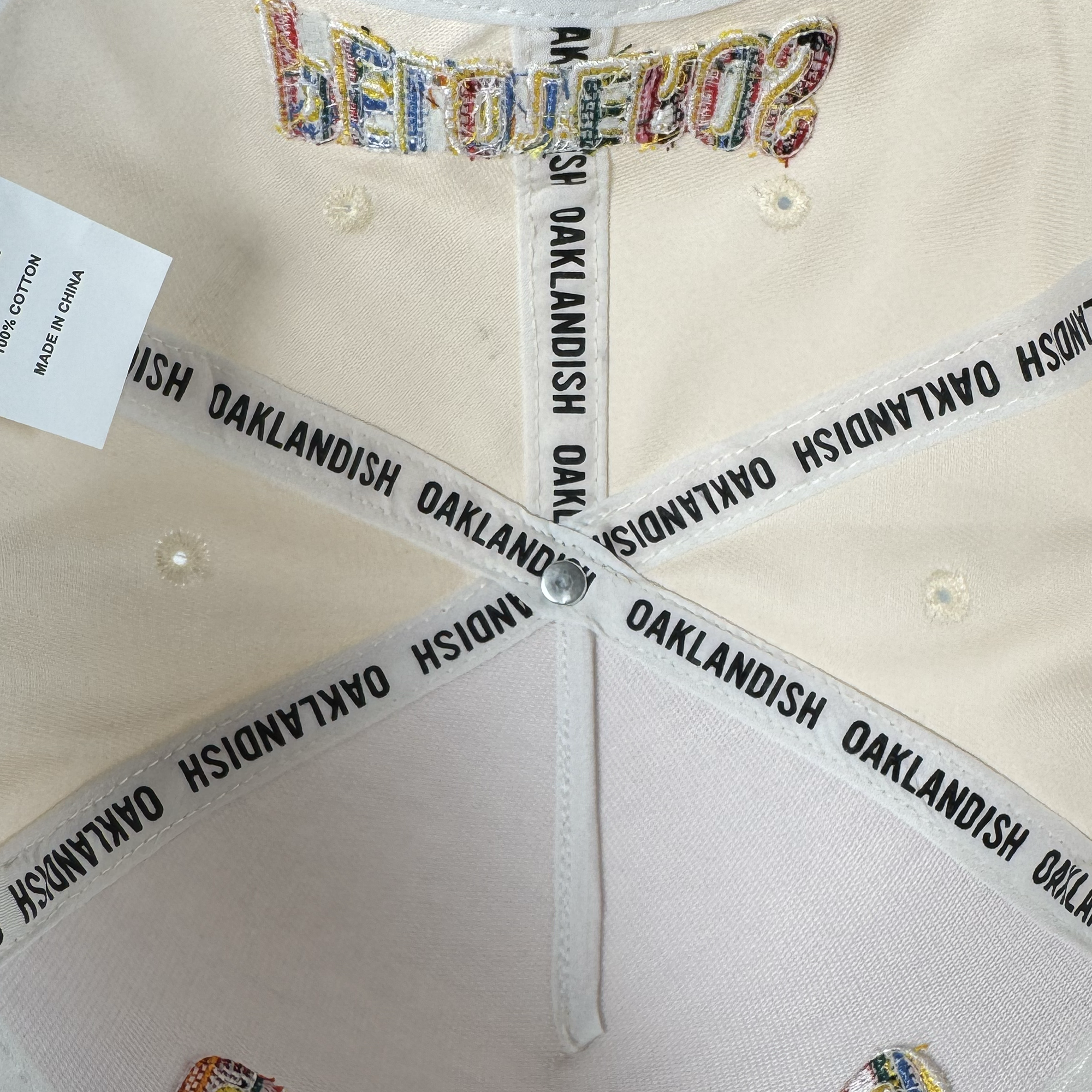 Close-up of the Oakland Beisbol 5 Panel Cap by Oakland Ballers reveals stitched labels, a "100% COTTON MADE IN CHINA" tag, and multicolored embroidery on the front panel nodding to the local baseball team.