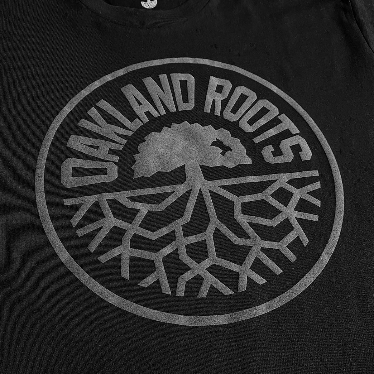 The Oakland Roots SC Blackout Logo Tee is a black shirt showcasing the Oakland Roots SC logo in gray. The design highlights a tree with sprawling roots extending downwards, enclosed in a circle, with "OAKLAND ROOTS" arcing over the top of the circle. Embrace your Oaklandish spirit and support your favorite sports club with this iconic tee.