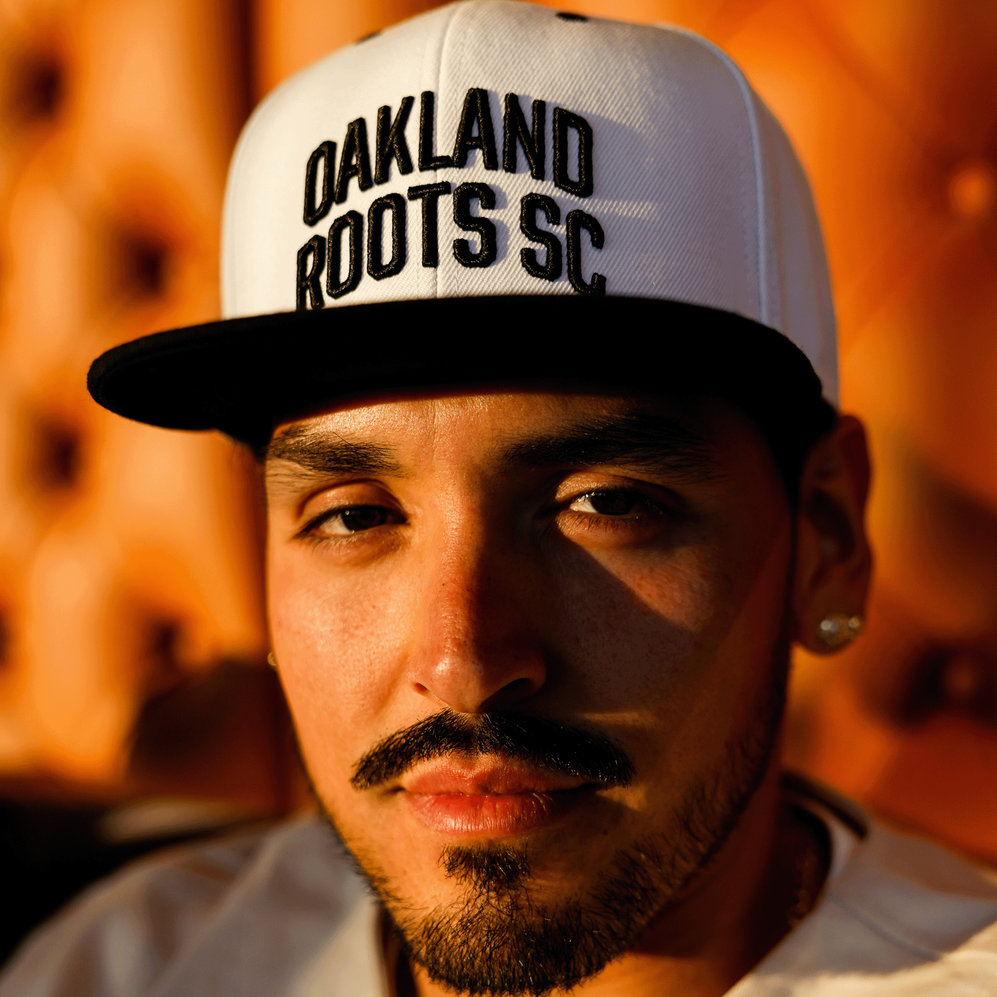 A man with a goatee and mustache wears the Oakland Roots SC Arched Text Snapback, featuring "OAKLAND ROOTS SC" written in black letters on a white hat. The background is softly blurred with warm tones, emphasizing the man's face illuminated by natural sunlight. He has a neutral expression.